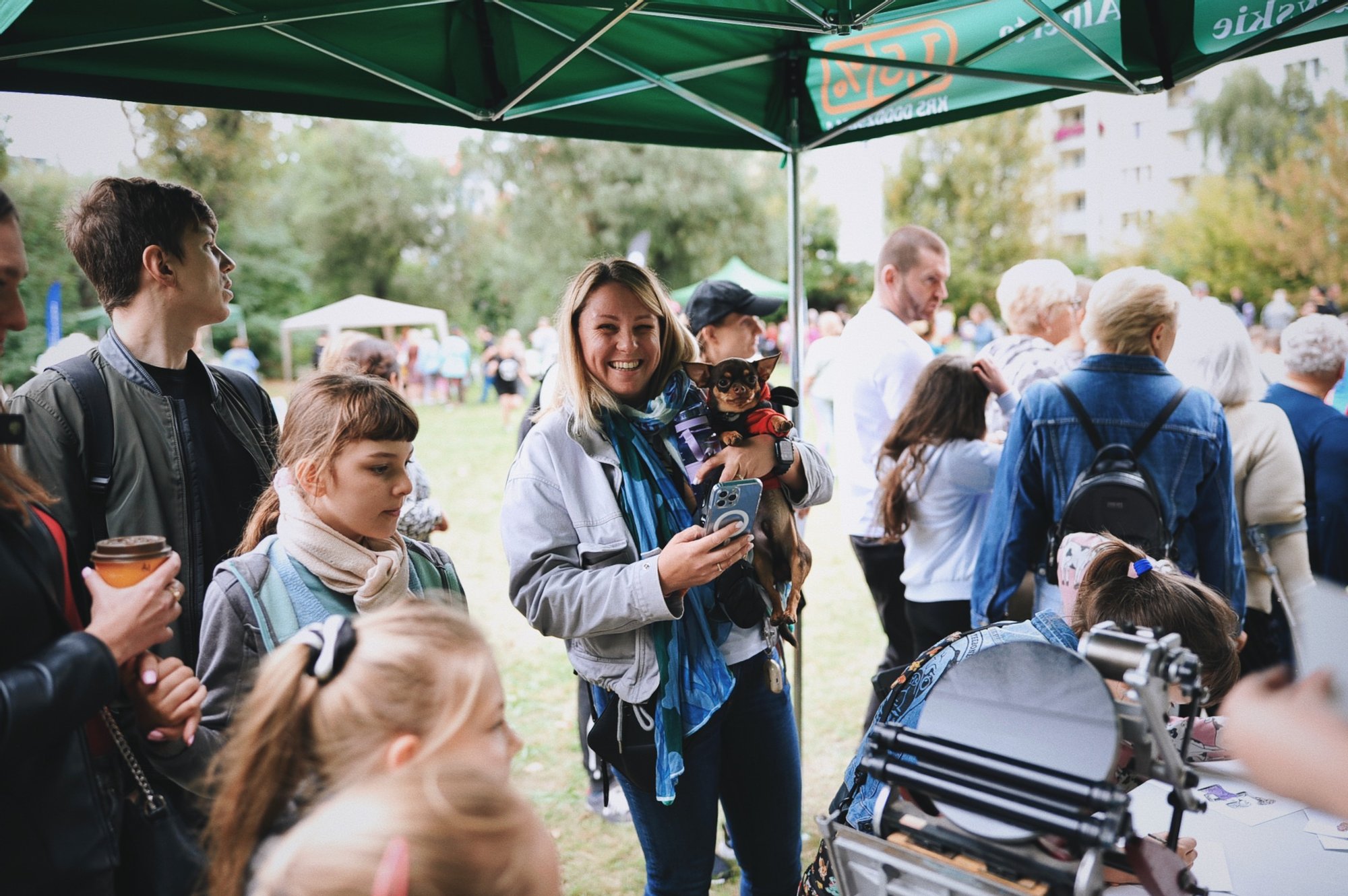 Kolorowa fotografia wykonana podczas osiedlowego pikniku. Pod dużym namiotem rozstawionym na łące stoi grupa osób w różnym wieku. Pośrodku stoi młoda kobieta z małym pieskiem na rękach. Uśmiecha się szeroko i patrzy w aparat.