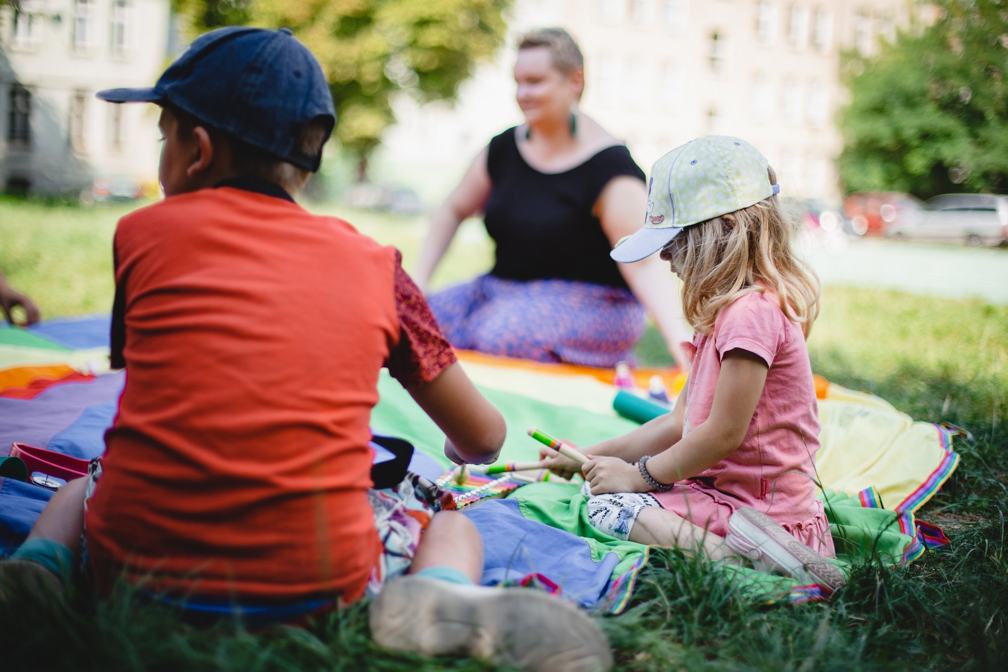 Grupa dzieci siedzi na kolorowej chuście na trawie. Przed nimi rozłożone instrumenty muzyczne.