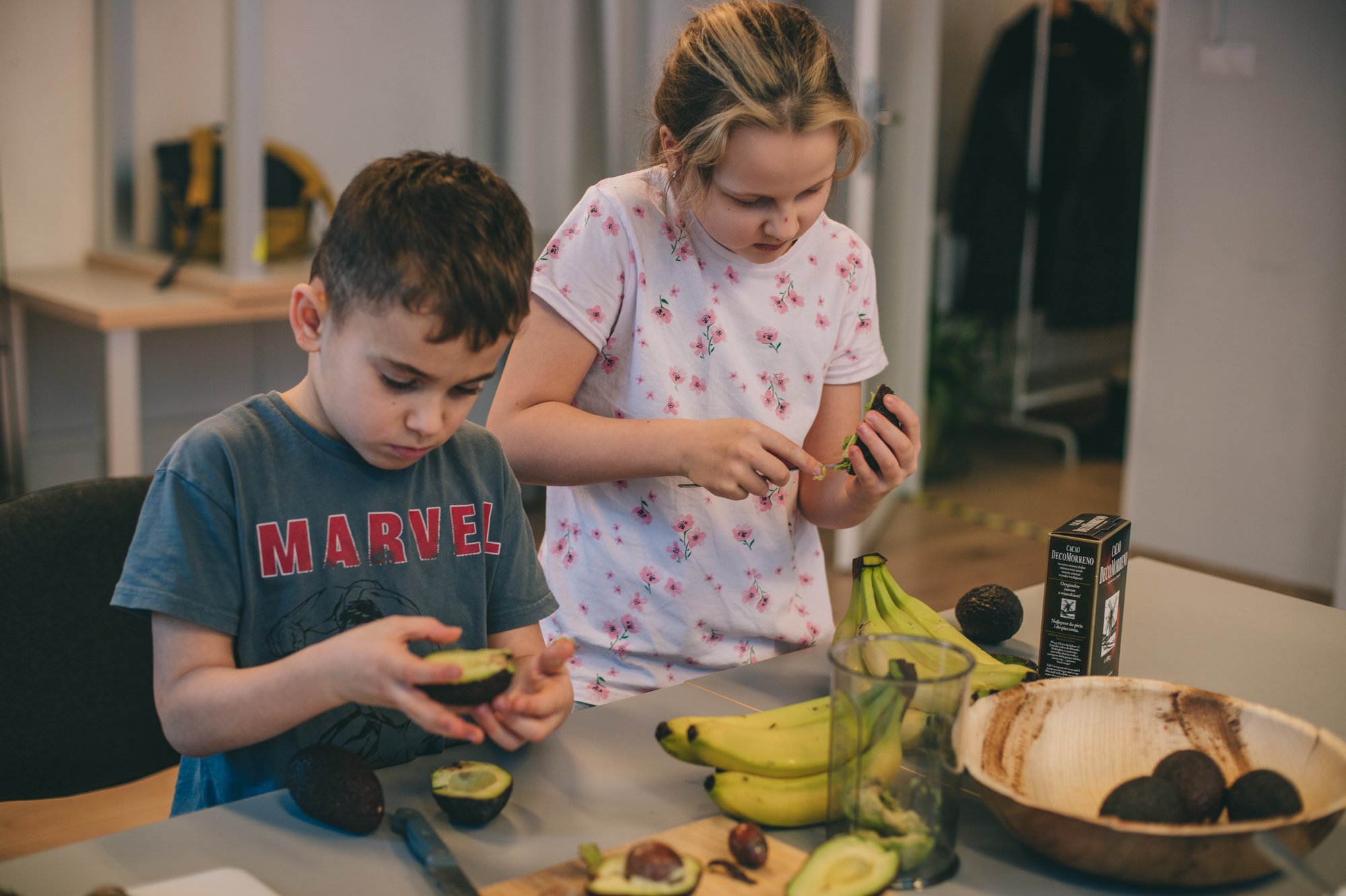 Kilkuletnie dzieci: chłopiec i dziewczynka stoją przy stole i wydrążają awokado. Przed nimi leżą jeszcze banany i kakao. Trwają zajęcia kulinarne.