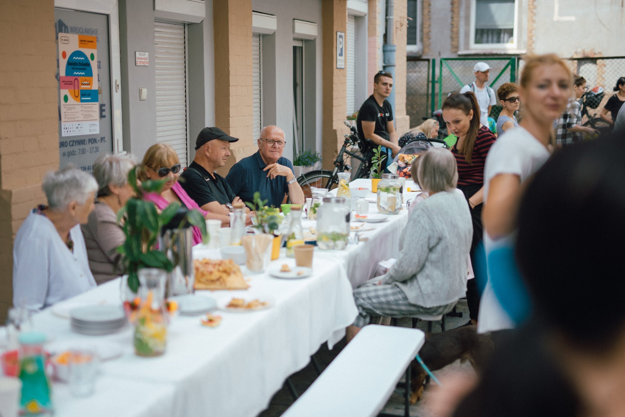 Przed budynkiem Centrum Aktywności Lokalnej Plac Grunwaldzki OD NOWA stoi długi stół, przykryty białym obrusem. Przy stole siedzi duża grupa seniorów i seniorek. Rozmawiają ze sobą. Na stole jest dużo przekąsek oraz napojów.