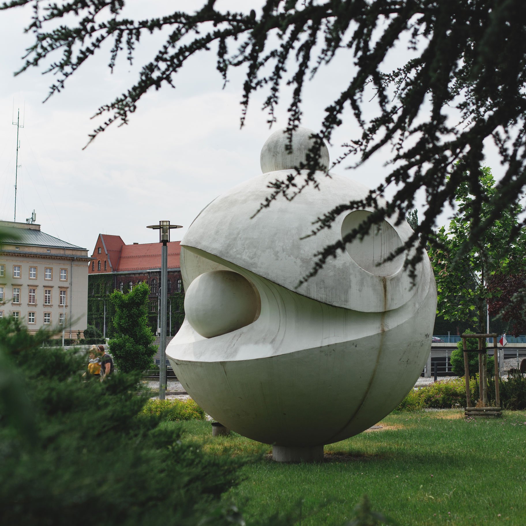 Na zdjęciu widać kamienną rzeźbę przedstawiająca model atomu, w tle widać budynki Muzeum Narodowego oraz Urzędu Wojewódzkiego. Na pierwszym planie widać rozmazane krzewy.