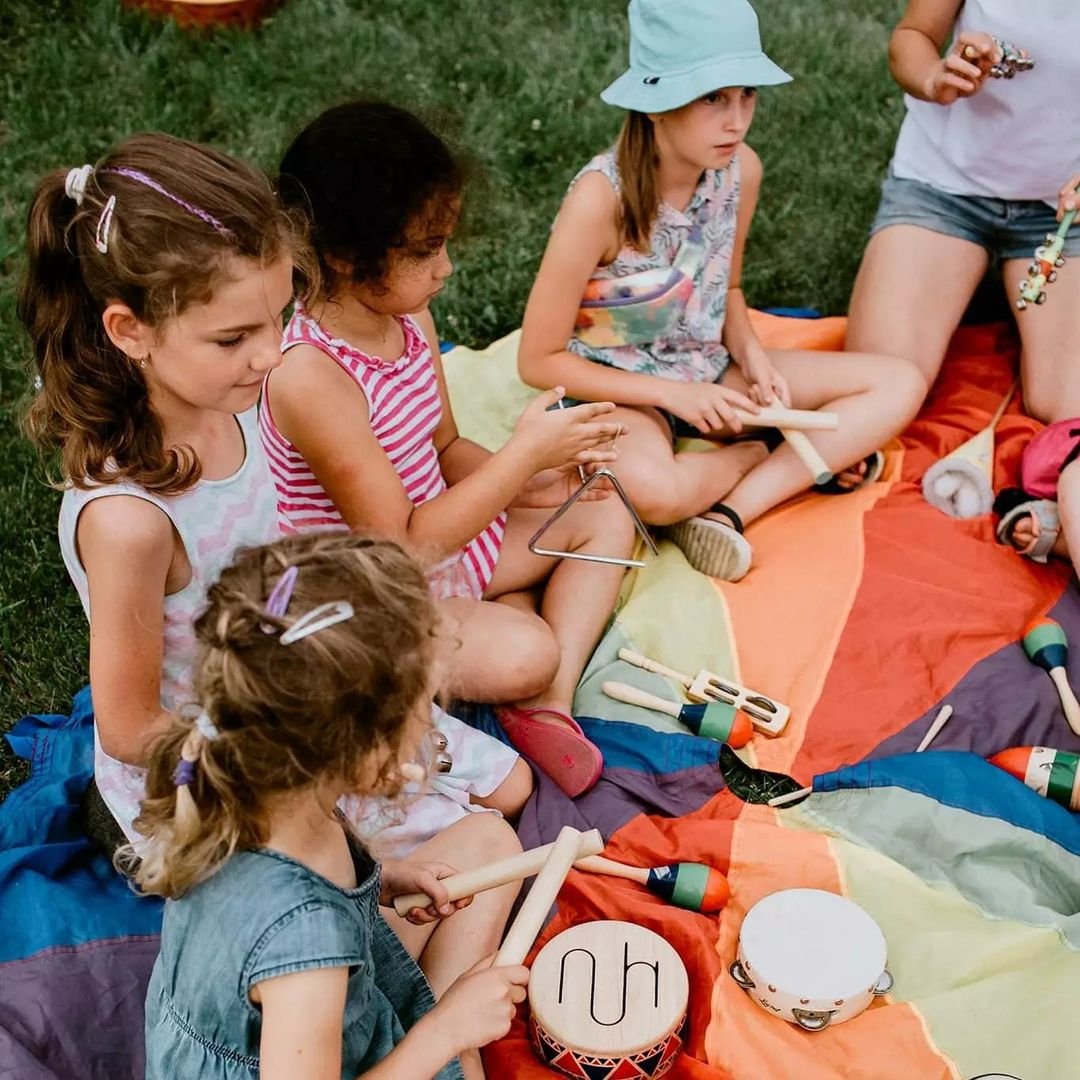 Na kolorowej chuście na trawie siedzi grupa dzieci. Przed nimi rozłożone instrumenty: tamburyn, trójkąt, cymbałki, bębenek, grzechotki.
