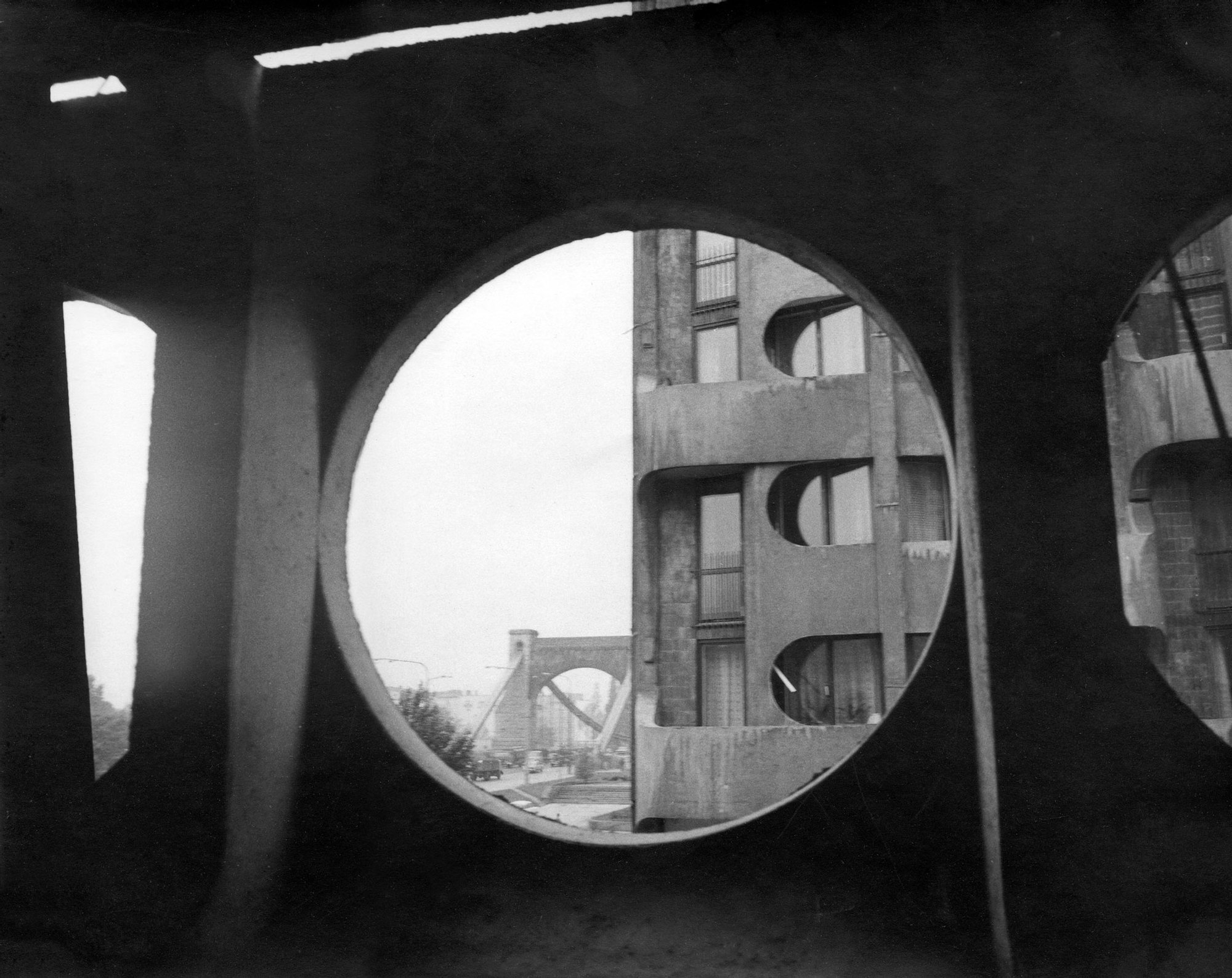 Stare, czarno-białe zdjęcie. Widok na część kompleksu bloków Manhattan i Most Grunwaldzki przez okrągłe okno z wewnątrz budynku.