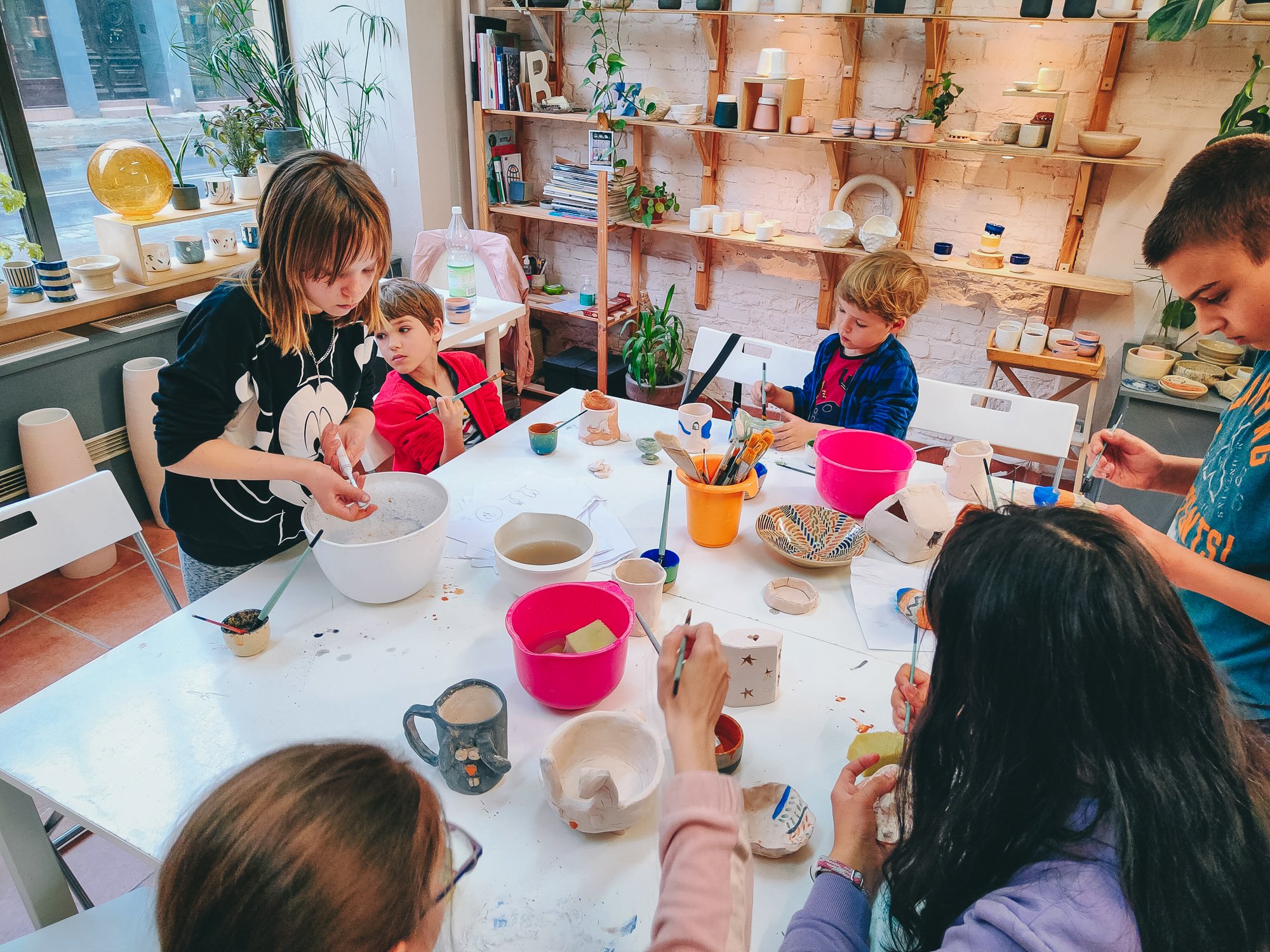 Grupa dzieci siedzi przy dużym stole w pracowni ceramicznej. Ozdabiają wcześniej ulepione przez siebie dzieła.