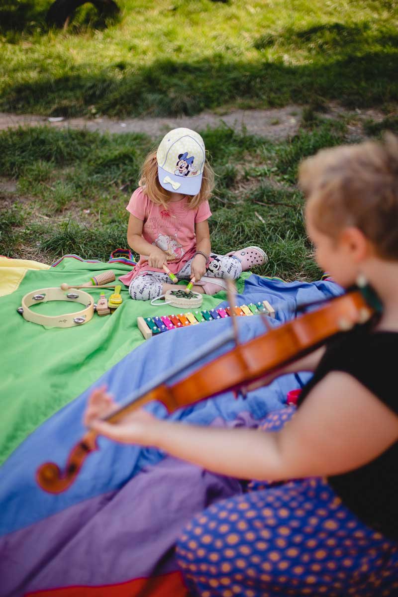 Na kolorowej chuście na trawie siedzi mała dziewczynka. Przed nią rozłożone instrumenty: tamburyn, cymbałki i bębenek.Obok niej siedzi kobieta grająca na skrzypcach.