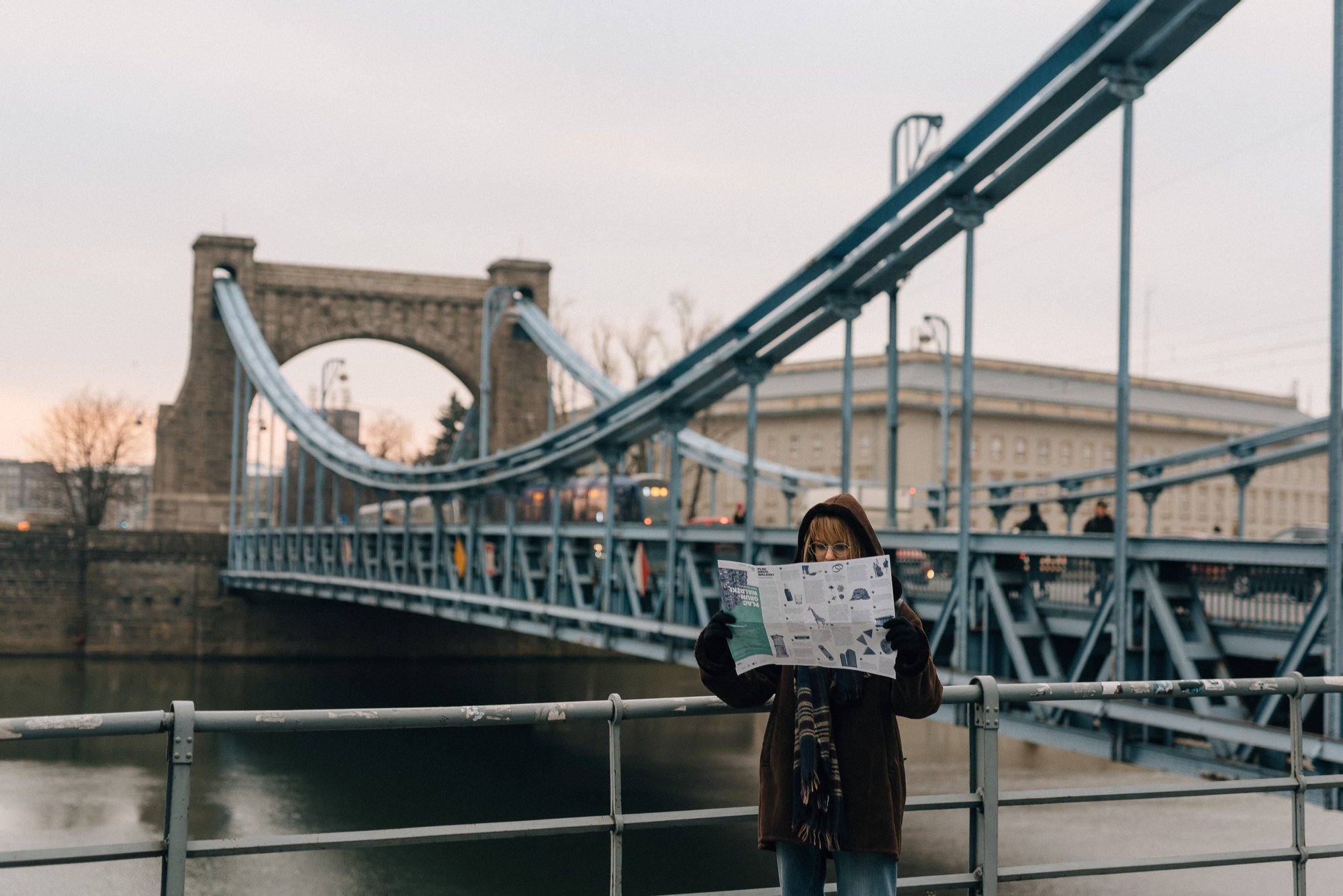 Dziewczyna w grubej kurtce, z kapturem na głowie i w rękawiczkach stoi na tle mostu Grunwaldzkiego i ogląda osiedlową mapę.