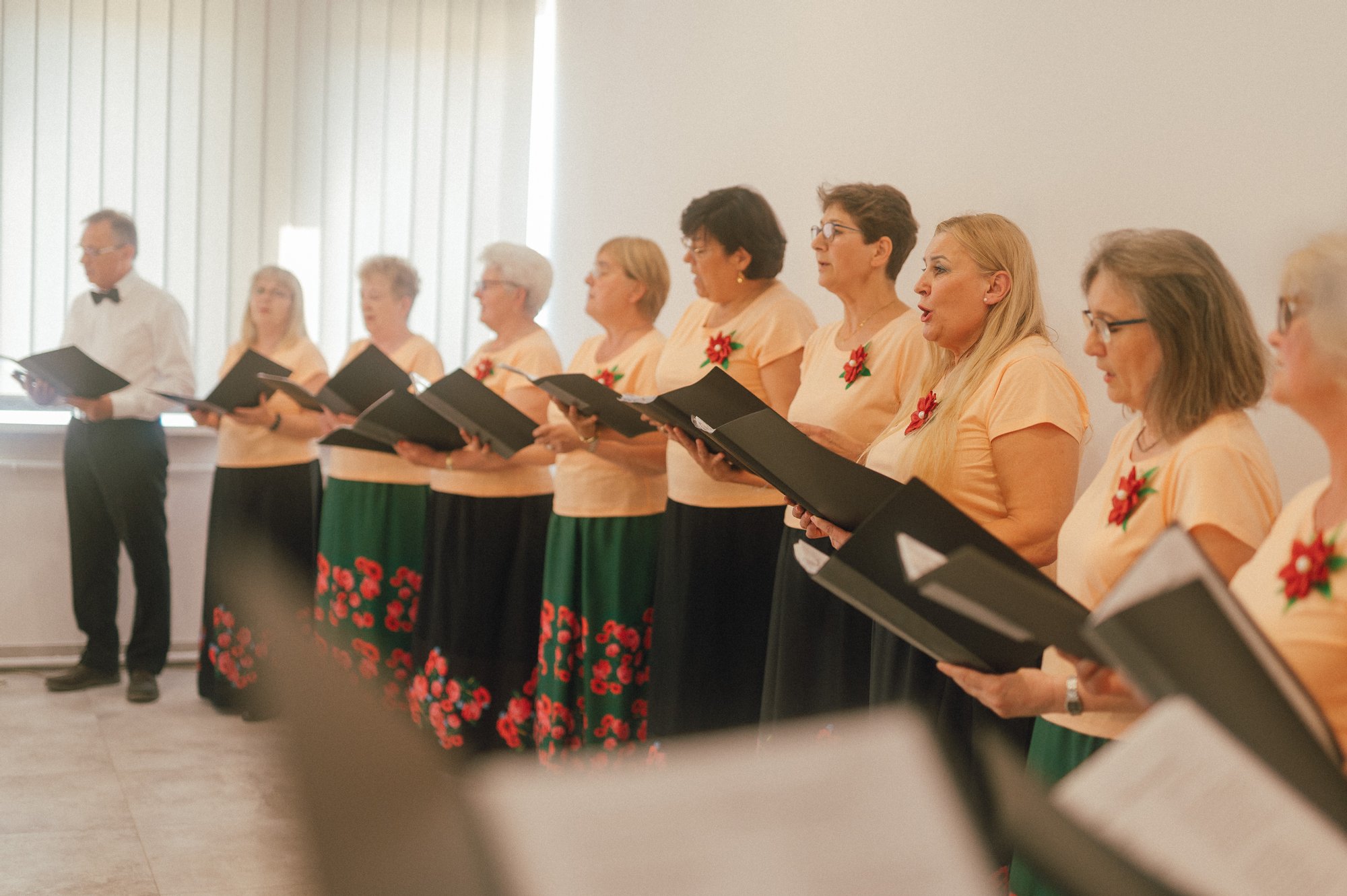 Kolorowa fotografia wykonana podczas występu chóru osiedlowego. W jasnym pomieszczeniu w rzędzie stoją chórzystki i jednen chórzysta.