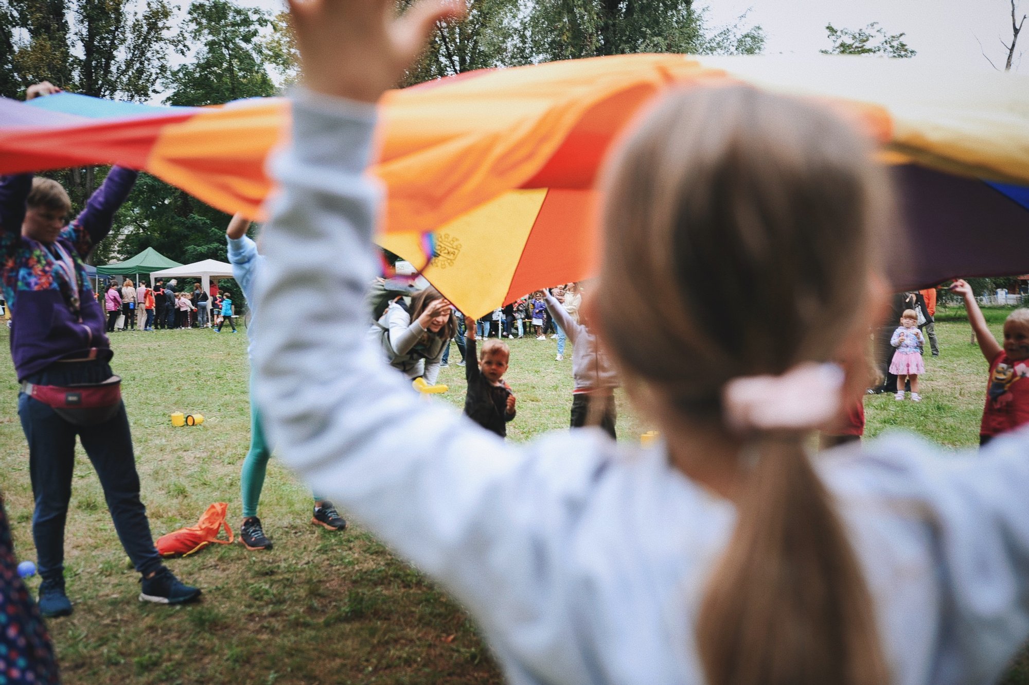 Kolorowa fotografia wykonana podczas osiedlowego pikniku. Grupa dzieci wraz z animatorkami bawią się kolorową chustą animacyjną.