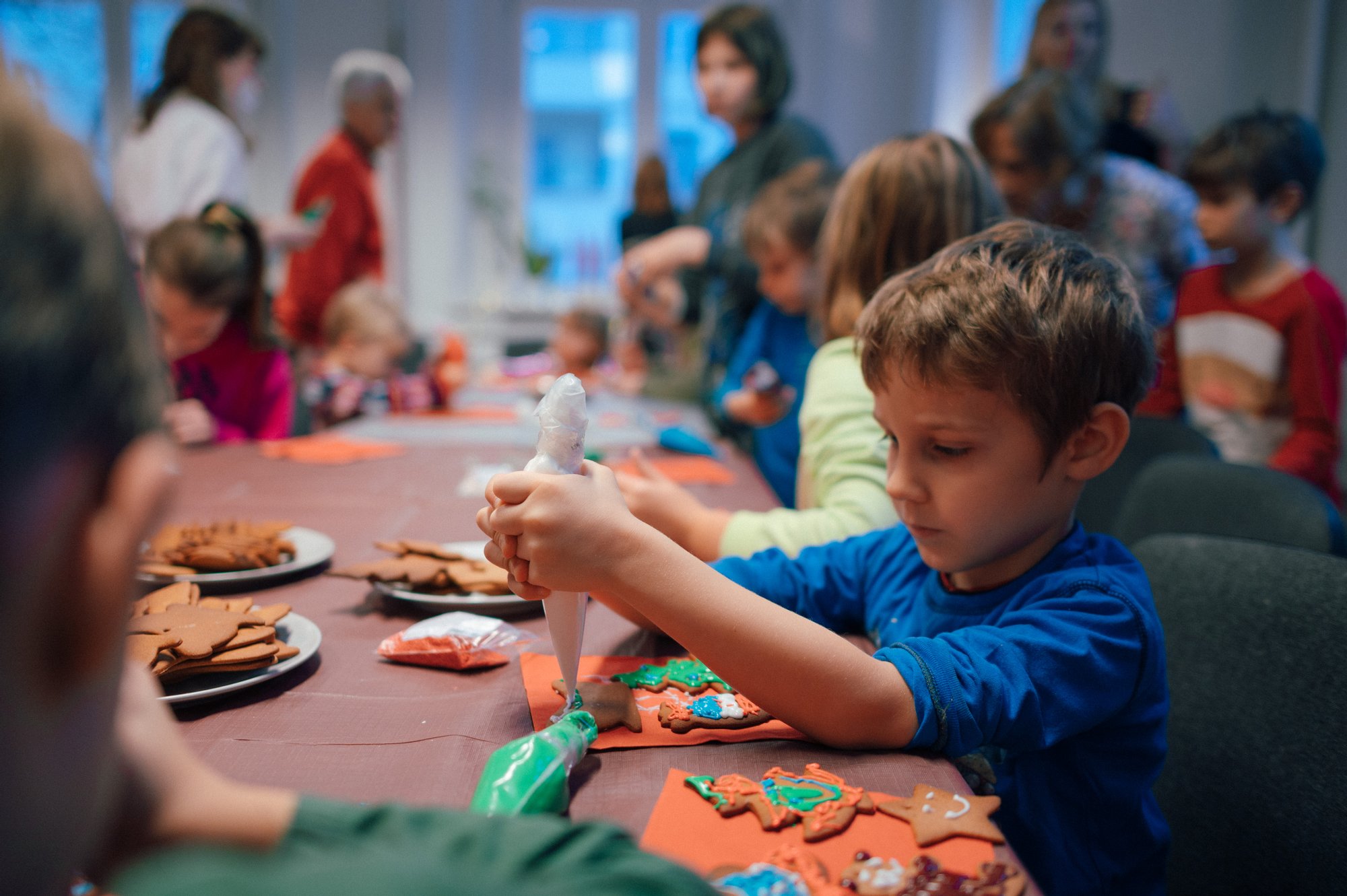 Kolorowa, pozioma fotografia. Na pierwszym planie kilkuletni chłopiec dekoruje lukrem ręcznie robione pierniczki. W tle inne dzieci ozdabiają swoje wypieki.