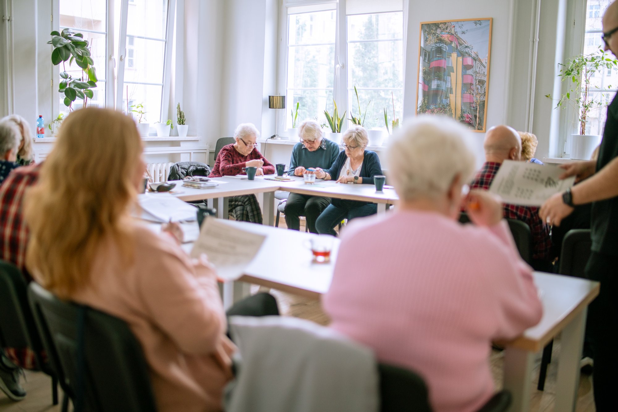 Kolorowa fotografia wykonana podczas warsztatów dla seniorów i seniorek. W dużym, jasnym pomieszczeniu przy długich stołach siedzą osoby starsze. Wykonują ćwiczenia pamięciowe i łamigłówki.