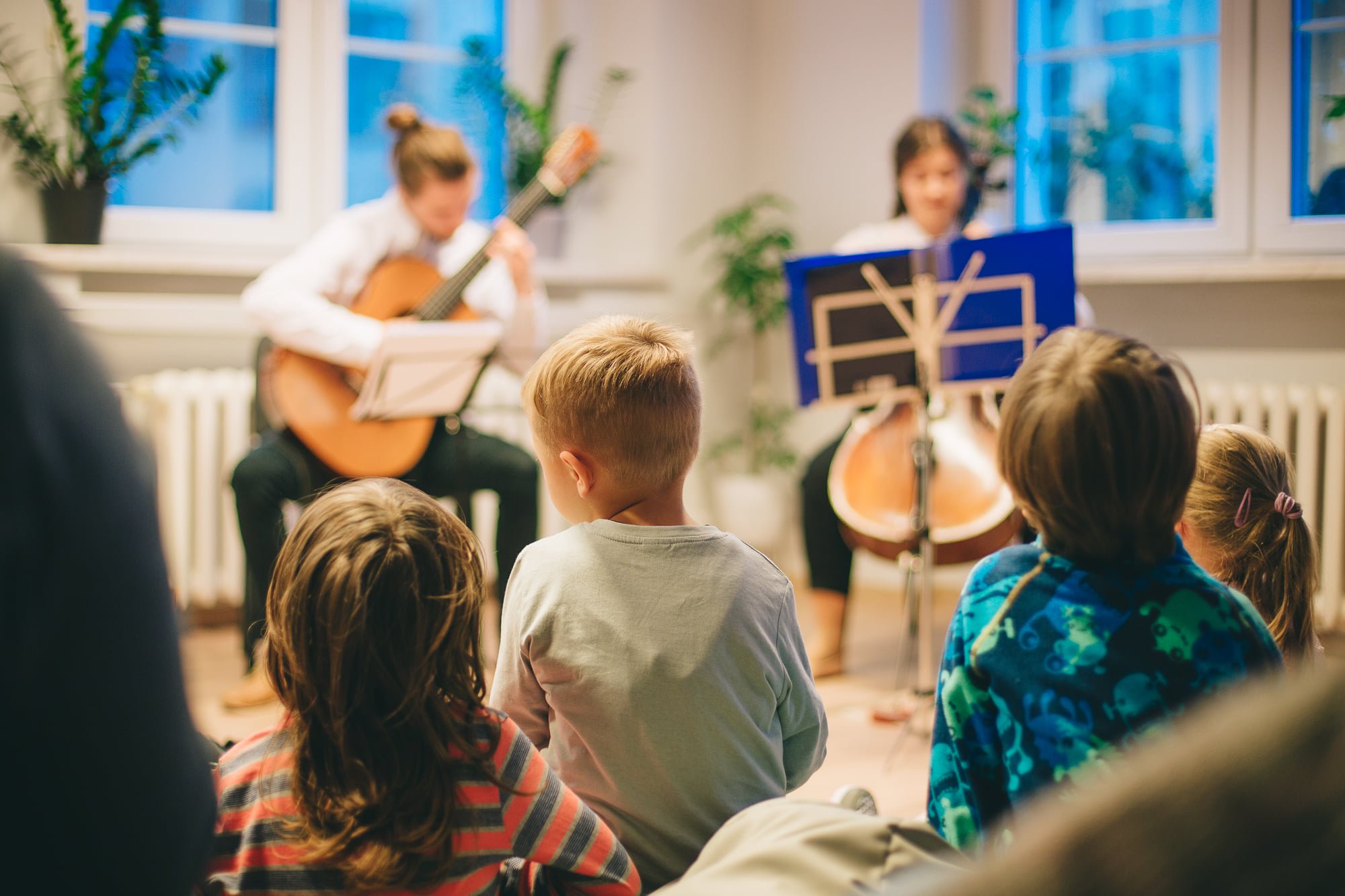 Na pierwszym planie zdjęcia widoczne dzieciaki zasłuchane w koncercie. W tle siedzą dwie osoby, grają na gitarze oraz wiolonczeli.