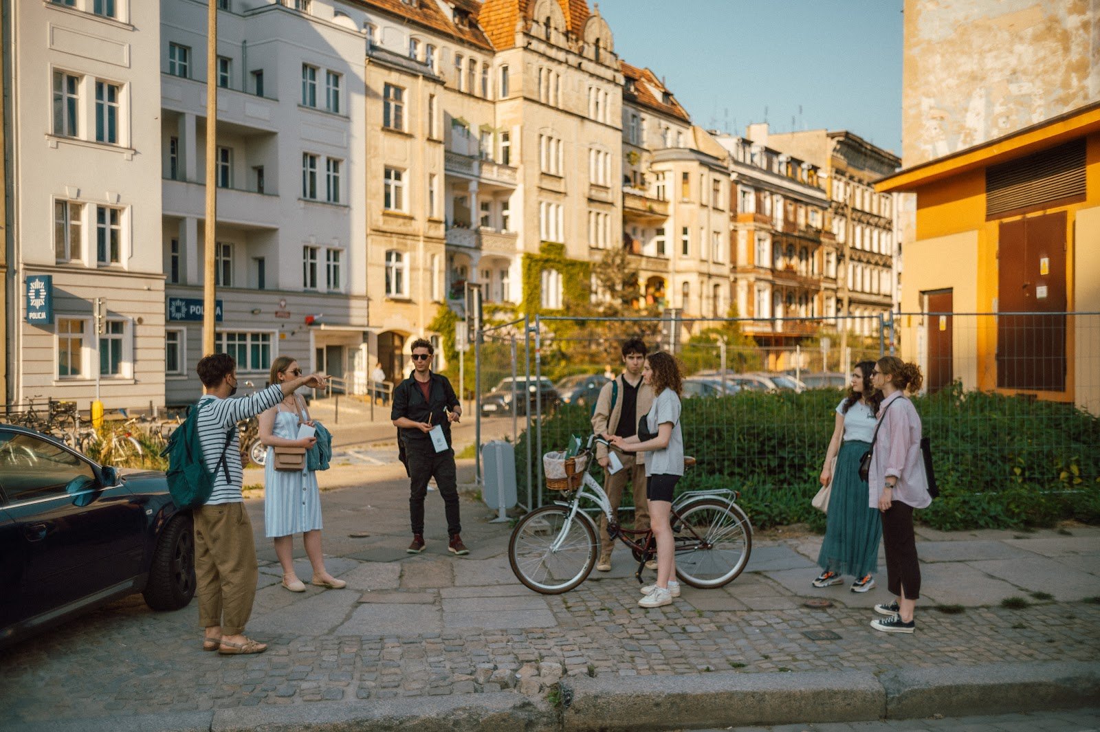 Grupa młodych ludzi stoi na chodniku i rozmawia. Jedna z osób trzyma rower, druga wskazuje ręką obiekt poza kadrem. 