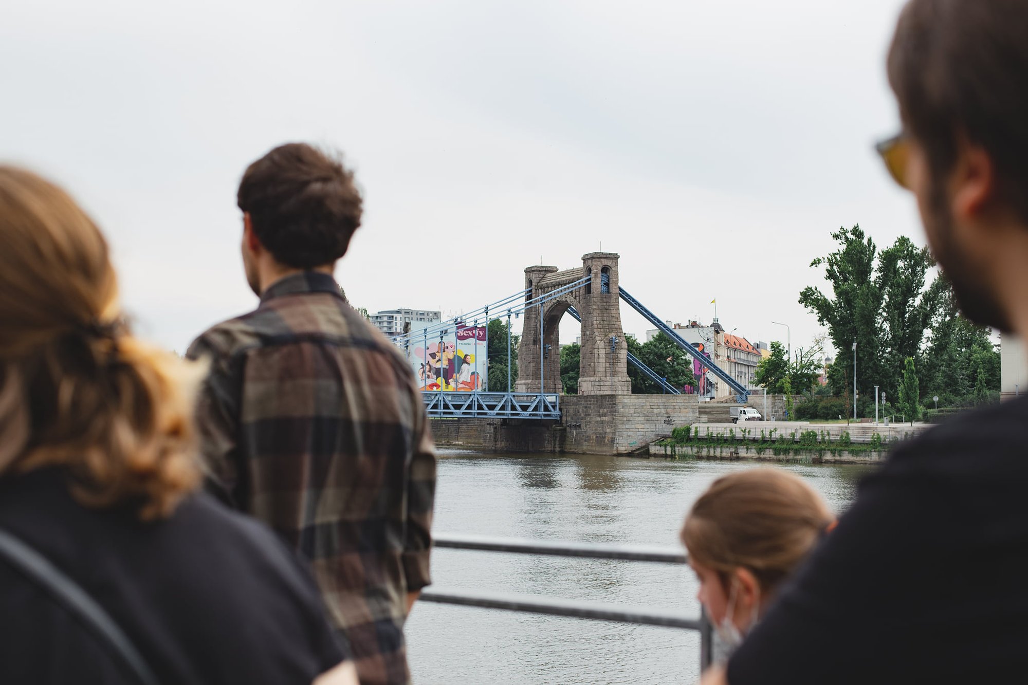 Kolorowa, pozioma fotografia przedstawiająca grupę osób podczas spaceru po osiedlu. Osoby spacerujące są rozmyte, ostrość zdjęcia ustawiona jest na most Grunwaldzki, znajdujący się w tle.