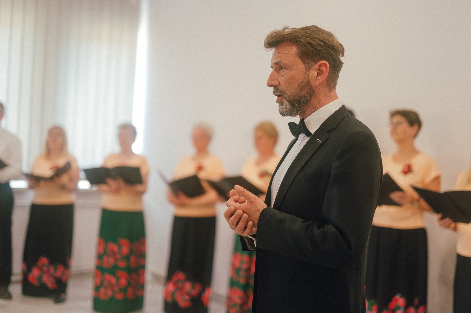 Kolorowa fotografia wykonana podczas występu chóru osiedlowego. Zbliżenie na eleganckiego mężczyznę w garniturze - to dyrygent. Za nim stojące w rzędzie chórzystki.