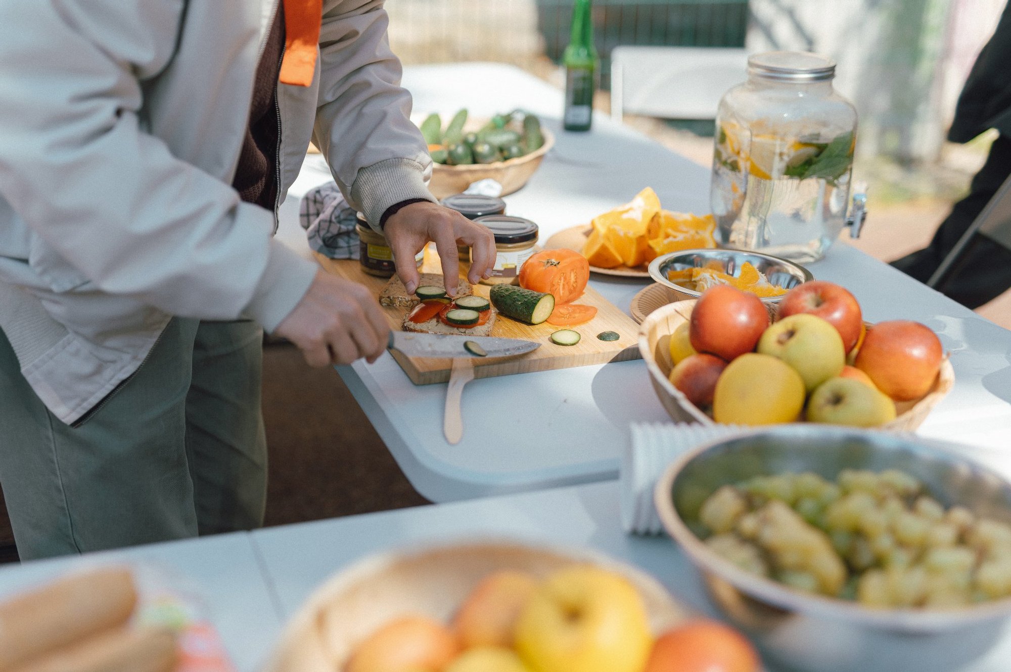 Na stołach rozstawionych na świeżym powietrzu stoi dużo owoców, warzyw oraz ciast. Przy stole stoją ludzie w różnym wieku i przygotowują potrawy.