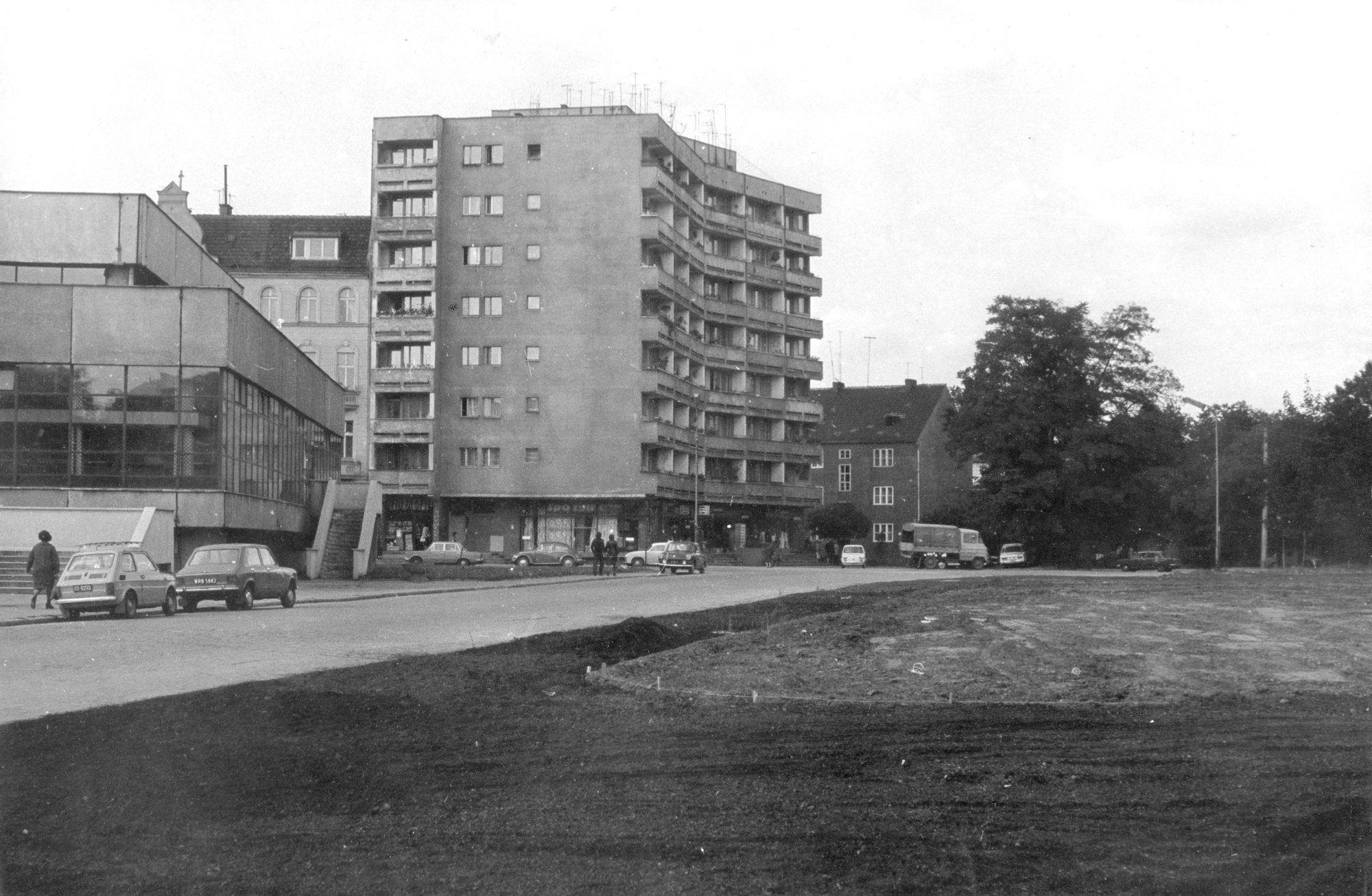 Czarno-biała archiwalna fotografia przedstawia budynki i samochody po lewej stronie, po prawej stronie rozkopana ziemia.