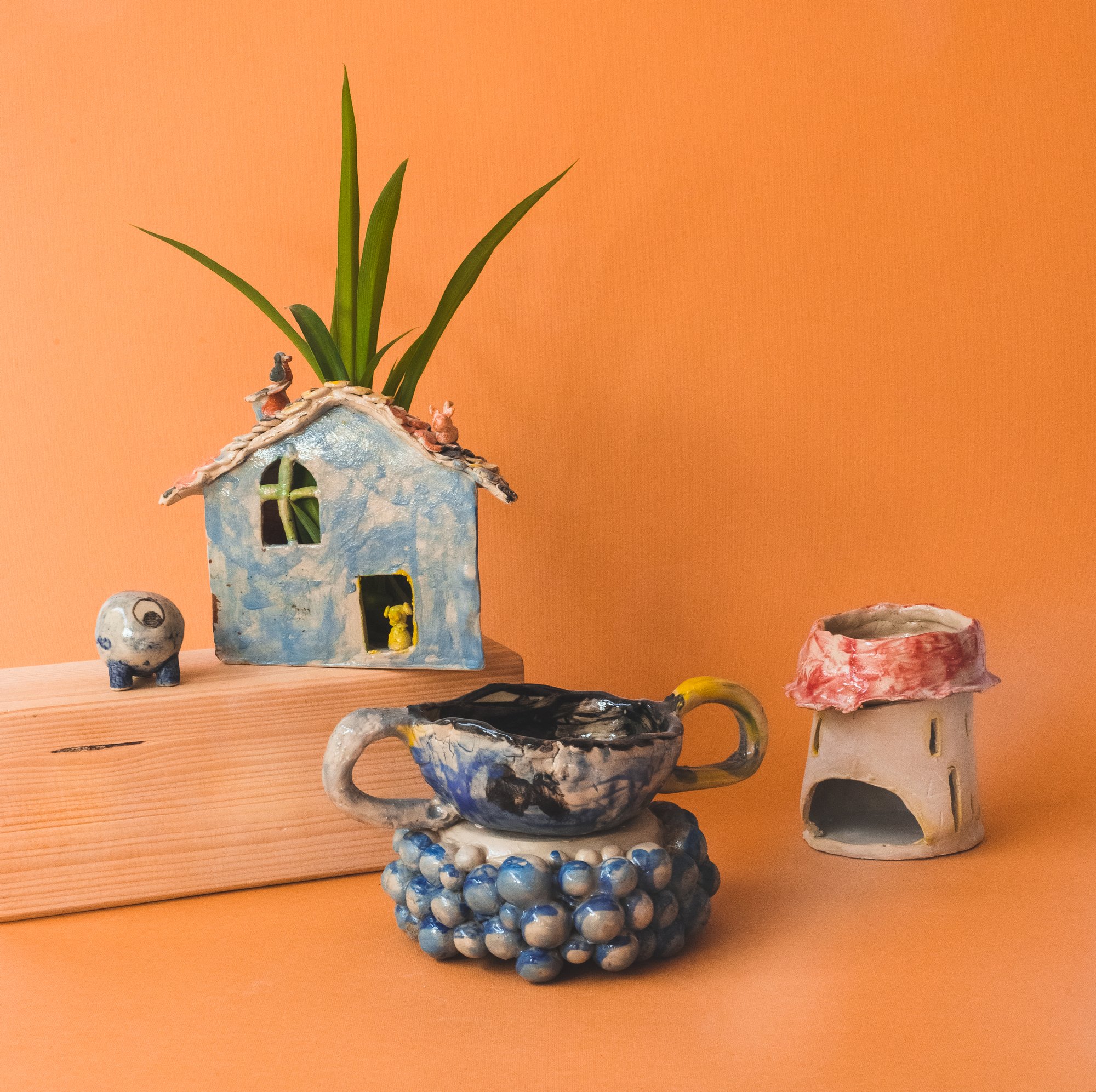 Na pomarańczowym tle zaprezentowane obiekty z ceramiki: domek-doniczka, kominek na świeczkę czy miseczka z dwoma uszkami.
