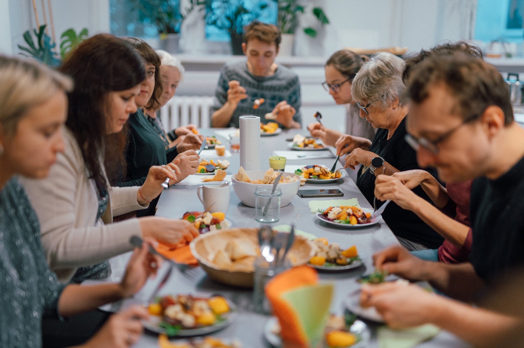 Kolorowa, pozioma fotografia. Grupa osób w różnym wieku siedzi przy długim stole zastawionym jedzeniem. To uczestnicy i uczestniczki warsztatów kulinarnych wspólnie jedzący to, co przygotowali podczas spotkania.