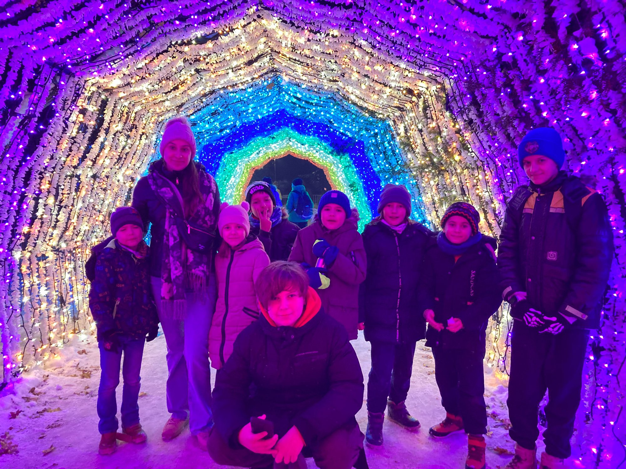 Kolorowa, pozioma fotografia: grupa dzieci w różnym wieku razem z opiekunką pozują do zdjęcia w tunelu zrobionym ze świątecznych, kolorowych światełek.
