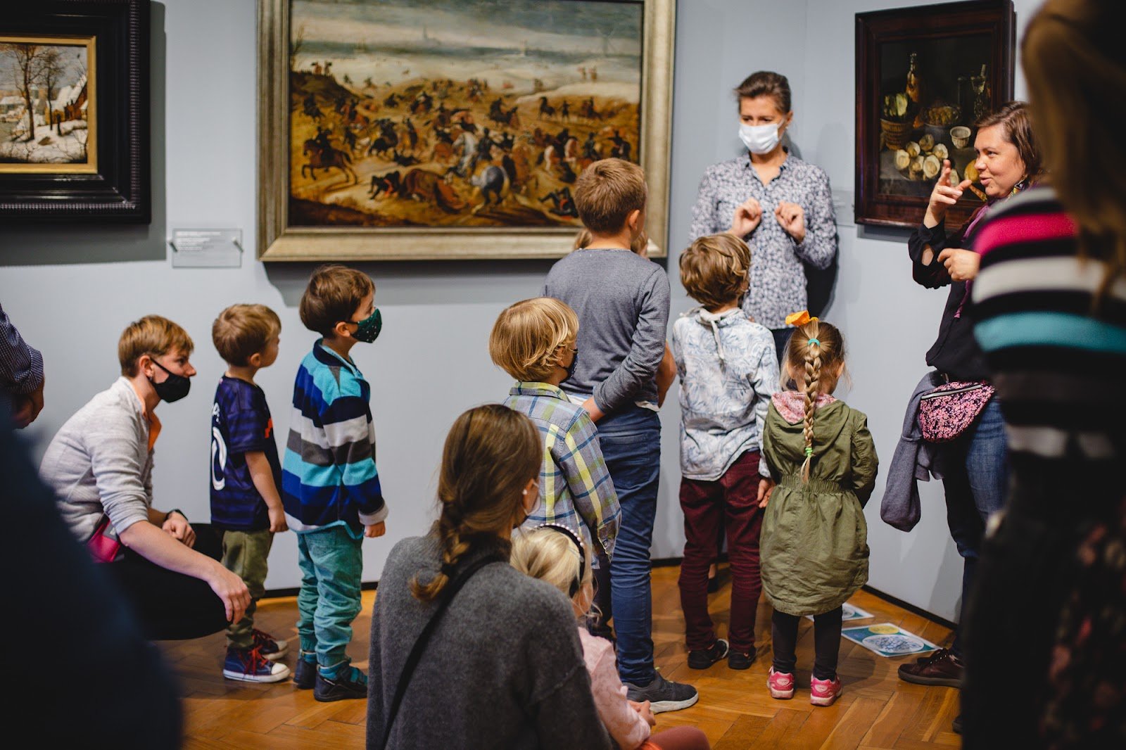 Grupa dzieci stoi grupą w muzeum, oglądają obrazy i słuchają co opowiada im przewodniczka, stojąca obok.