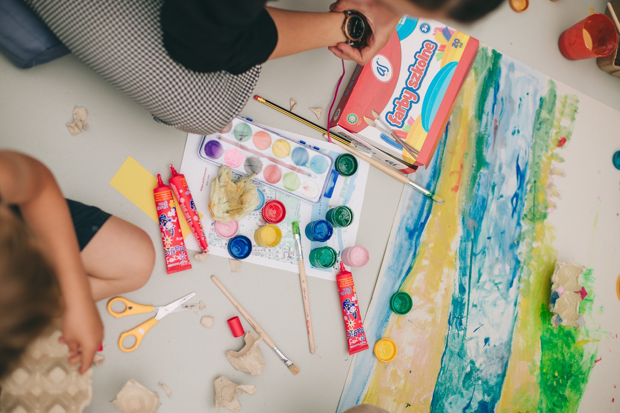 Dwójka dzieciaków siedzi na podłodze i maluje farbami. Przed nimi oprócz obrazu leżą farby, kleje, pędzelki i różne materiały plastyczne.