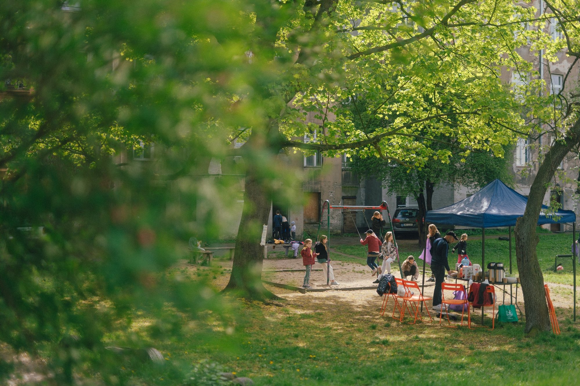 Kolorowa, pozioma fotografia przedstawiająca grupę dzieci i dorosłych spędzających czas na podwórku otoczonym kamienicami. 