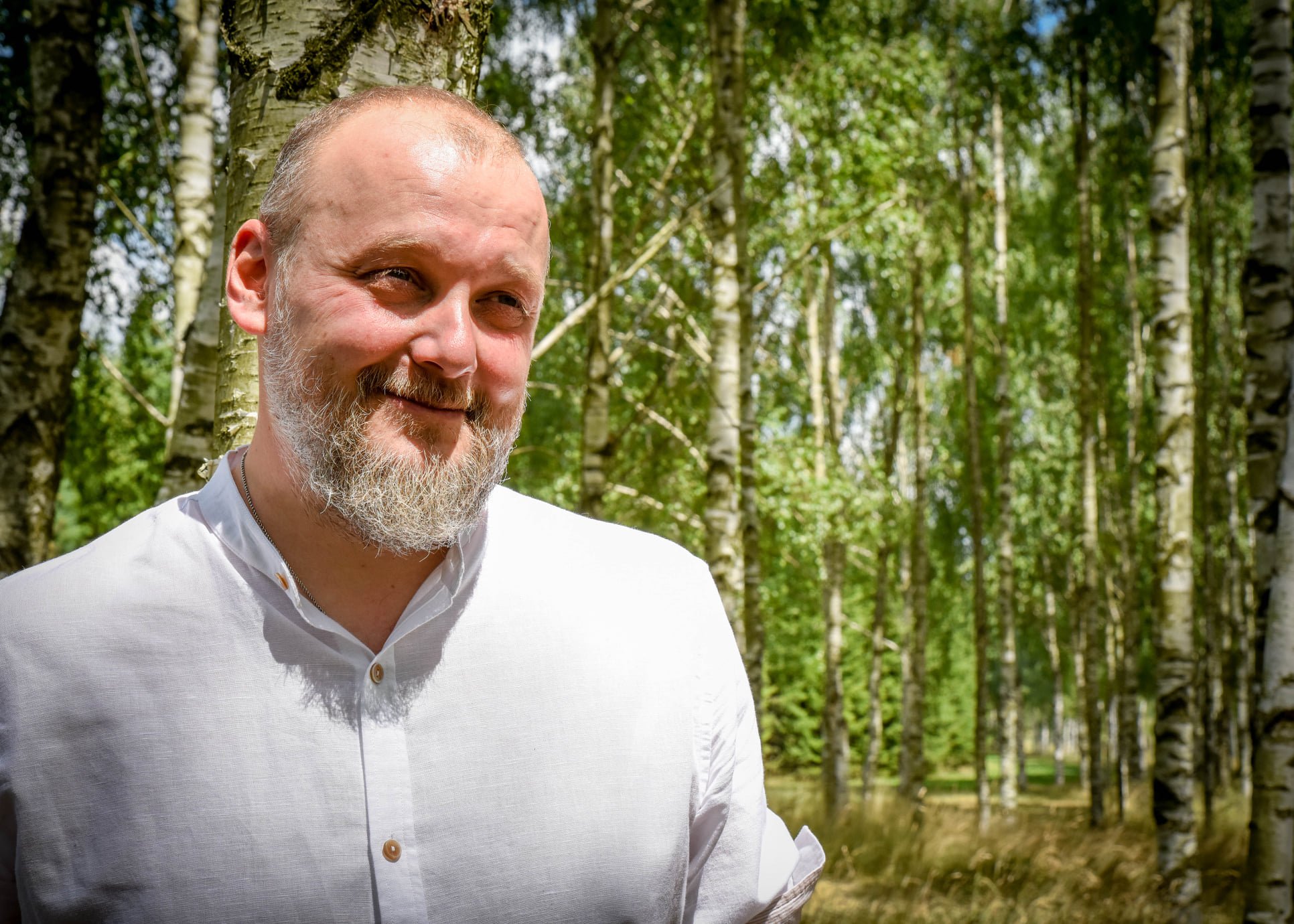 Uśmiechnięty mężczyzna z siwą brodą, ubrany w białą, zapinaną na guziki koszulę, stoi na tle brzozowego lasu.