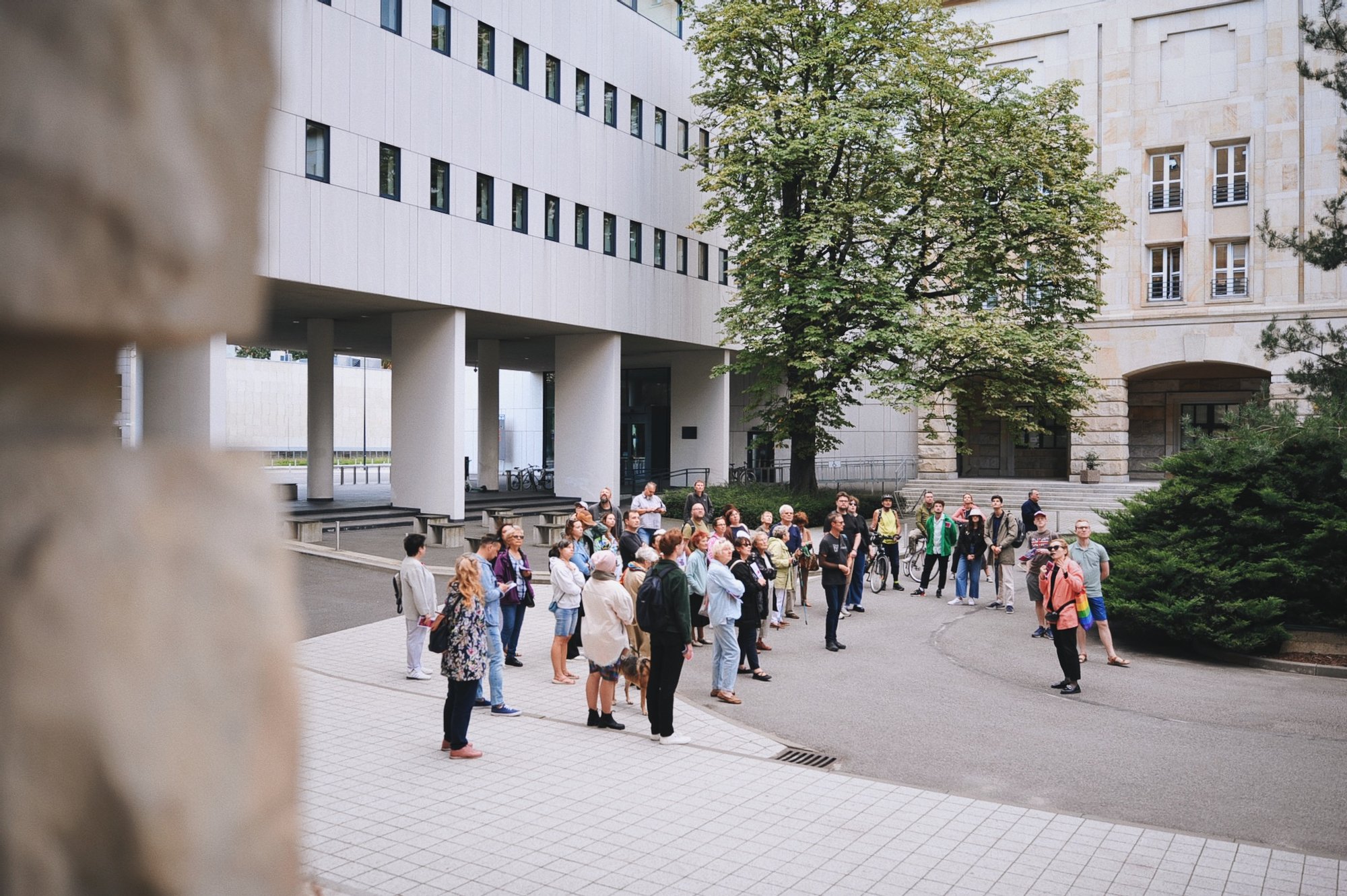 Kolorowa, pozioma fotografia: grupa osób w różnym wieku zebrała się przy budynku biblioteki Politechniki Wrocławskiej. Trwa spacer po osiedlu. 