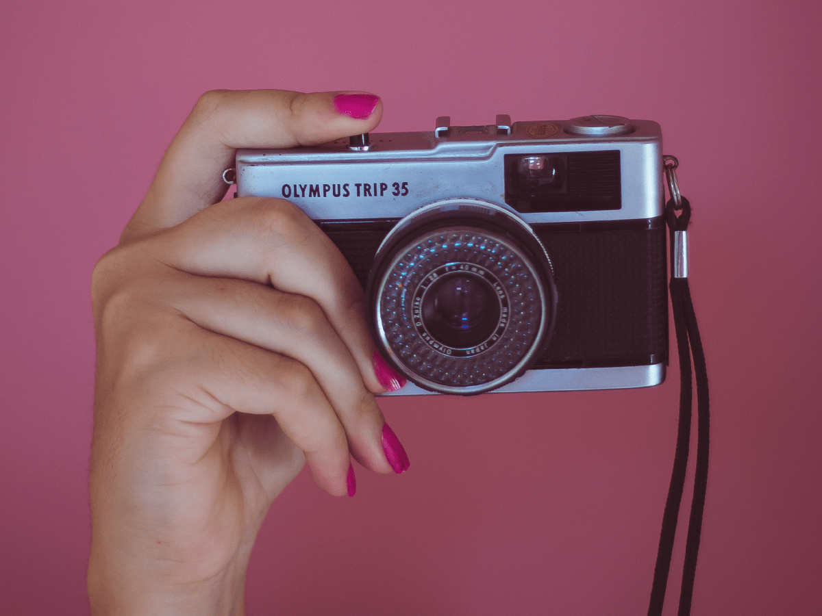 Zbliżenie na dłoń trzymającą stary aparat fotograficzny marki Olympus. Paznokcie dłoni pomalowane są na różowy kolor. Różowe jest również tło zdjęcia.