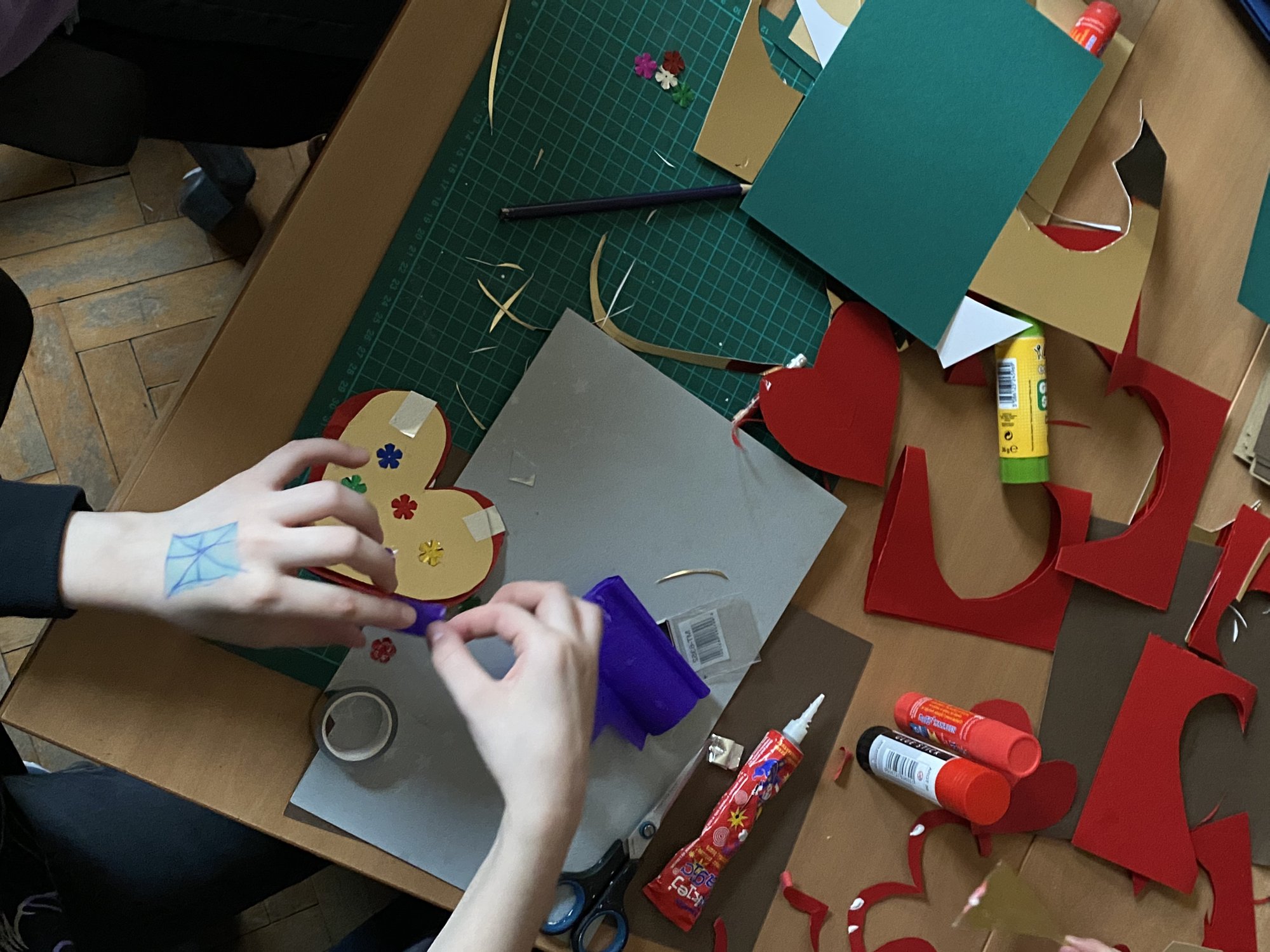 Zbliżenie na dziecięce dłonie, które wykonują ozdobną kartkę w kształcie serca. Na stole porozkładane kolorowe papiery, kleje i inne artykuly plastyczne.