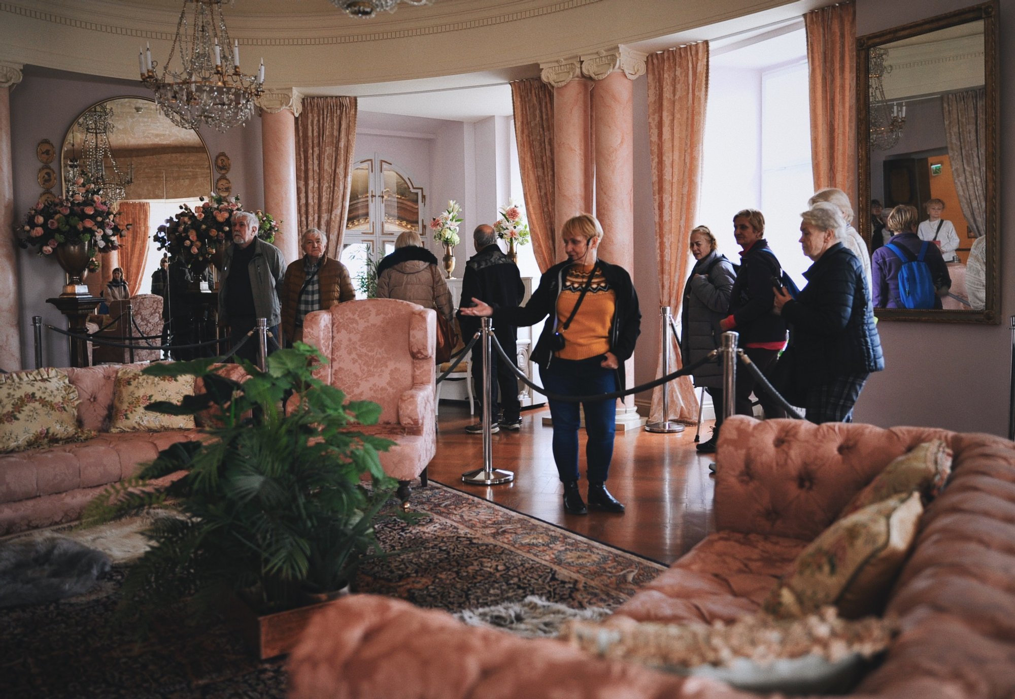 Kolorowa, pozioma fotografia. Grupa seniorów i seniorek zwiedza Zamek w Wałbrzychu. Na środku zdjęcia przewodniczka opowiada o meblach, które znajdują się  wobecnie oglądanym pomieszczeniu.