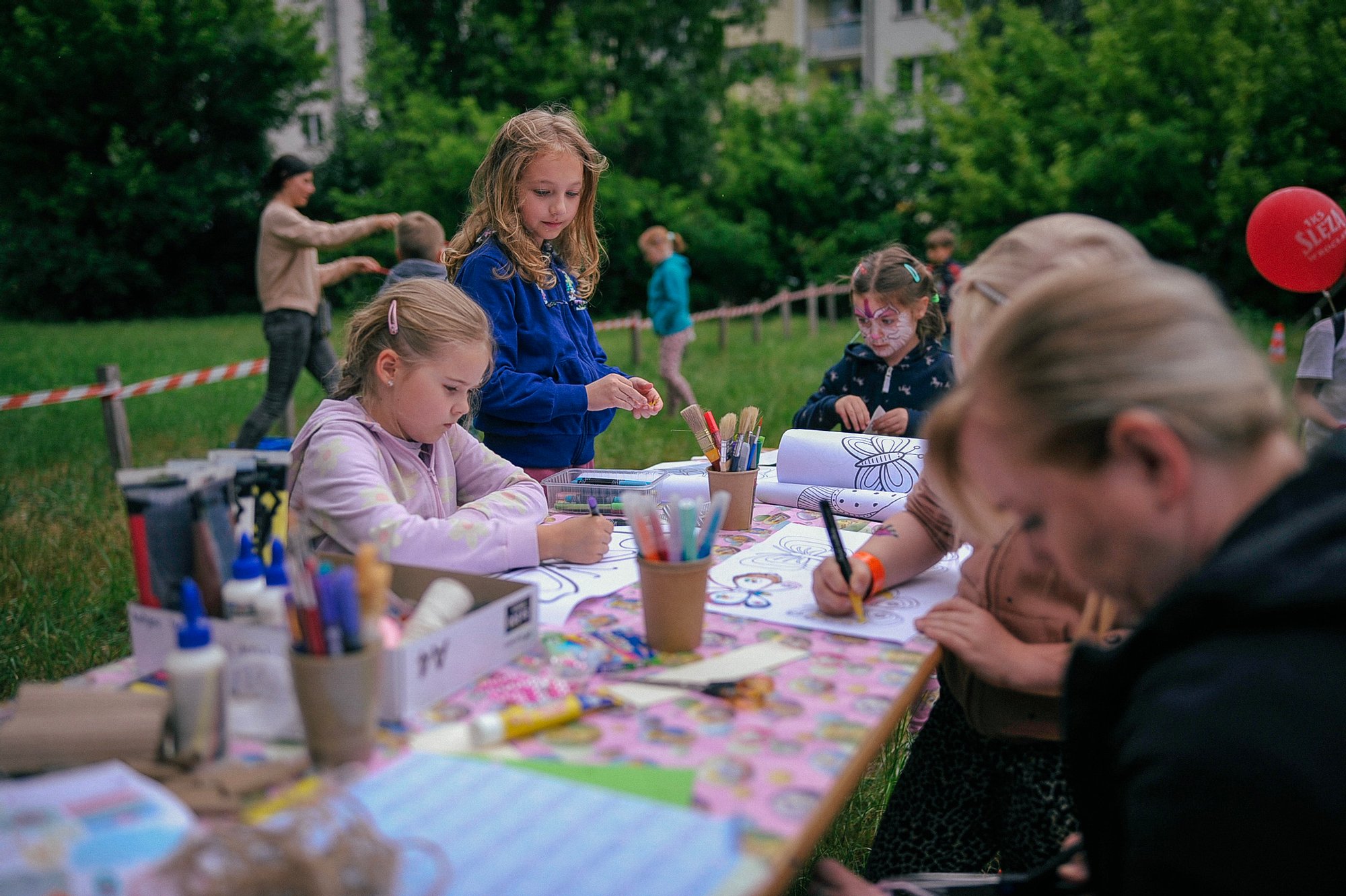 Kolorowa fotografia wykonana podczas święta osiedla. Przy długim stole rozstawionym na łące siedzą dzieci i zamjują się kolorowankami.