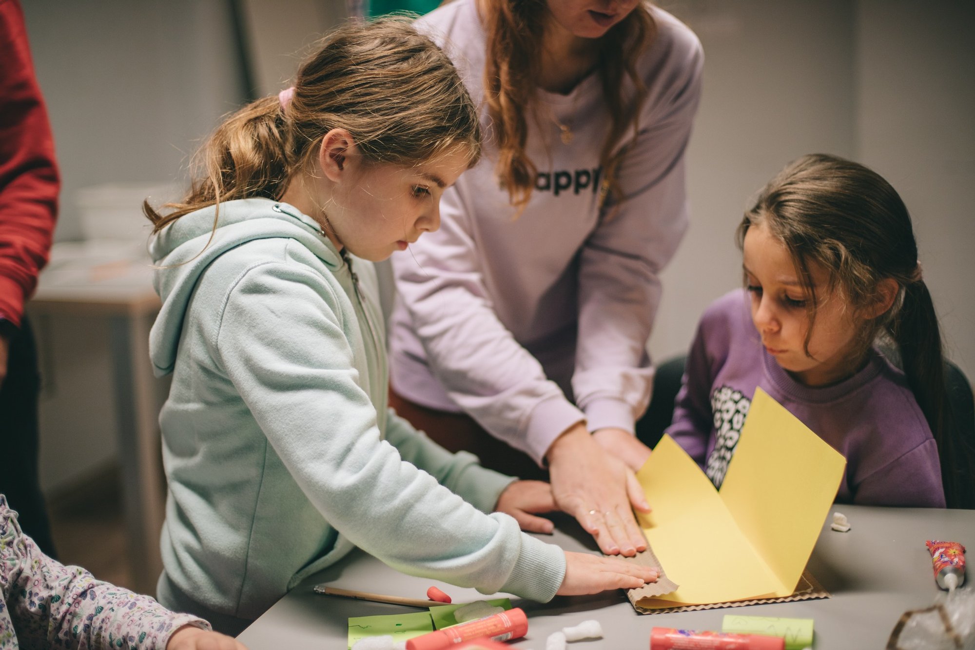 Dwie kilkuletnie dziewczynki wraz z prowadzącą zajęcia tworzą ozdoby z papieru przy dużym stole, na którym porozkładane są różne materiały papiernicze.