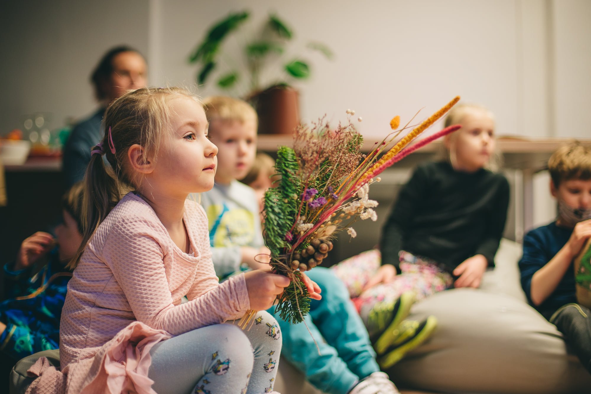 Grupa dzieci siedzi na poduszkach i z uwagą kogoś słuchają. Dziewczynka na pierwszym planie trzyma w rękach bukiet z suszonych kwiatków, gałązek i traw.
