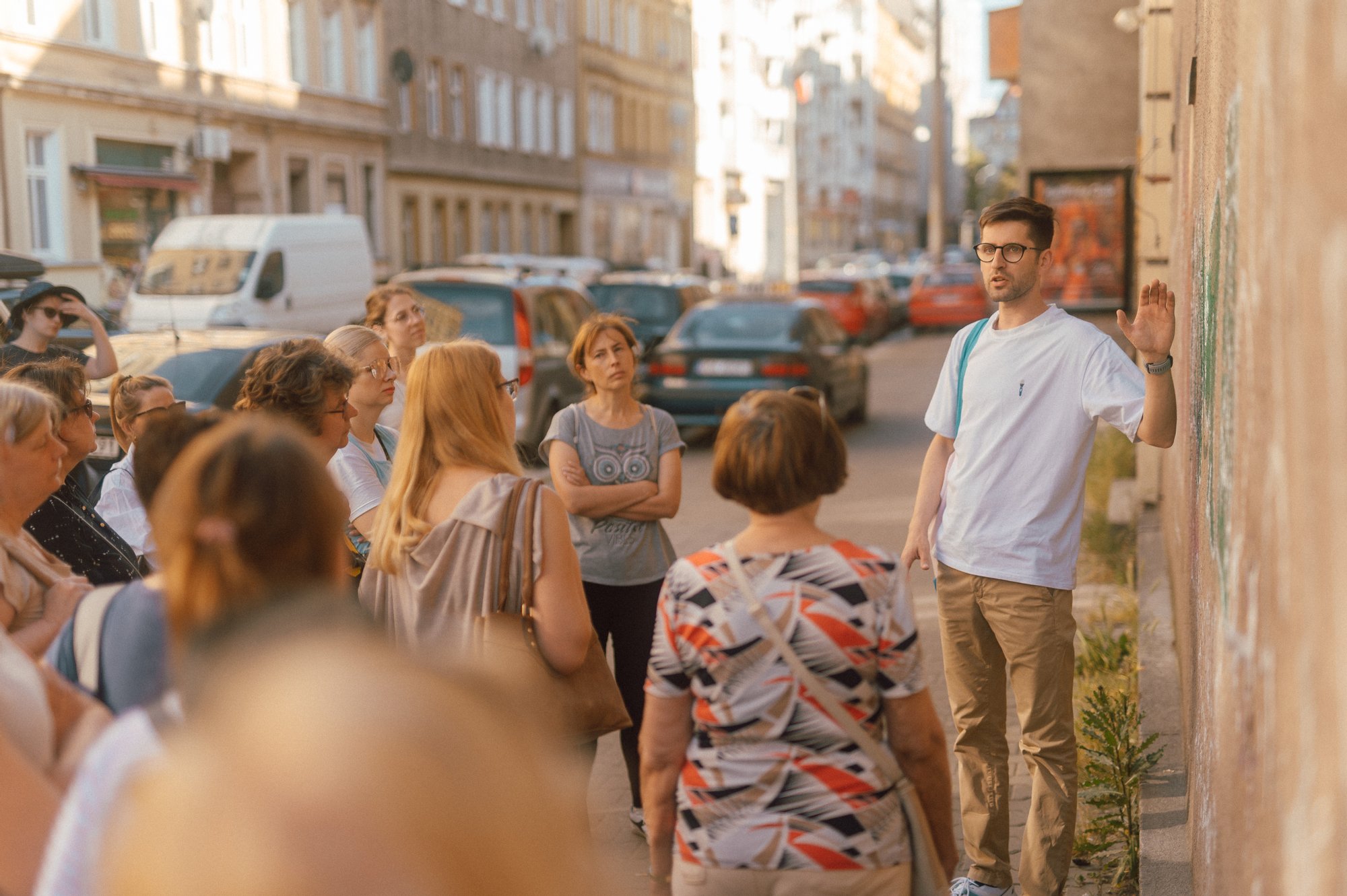 Kolorowa, pozioma fotografia, przedstawiająca grupę osób w różnym wieku, stojącą na chodniku. Przed nimi Maciek Wlazło - przewodnik spaceru, opowiadający o miejscu, w którym przystanęła grupa.