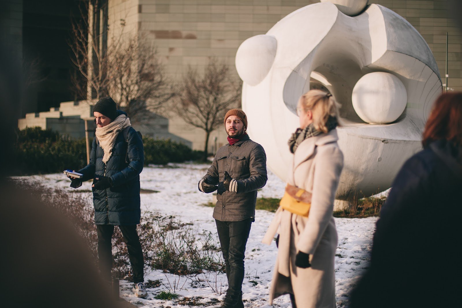 Grupa osób stoi pod rzeźbą atomu w zimowy dzień. Na ziemi widoczny śnieg, a ludzie poubierani są w ciepłe ubrania. 