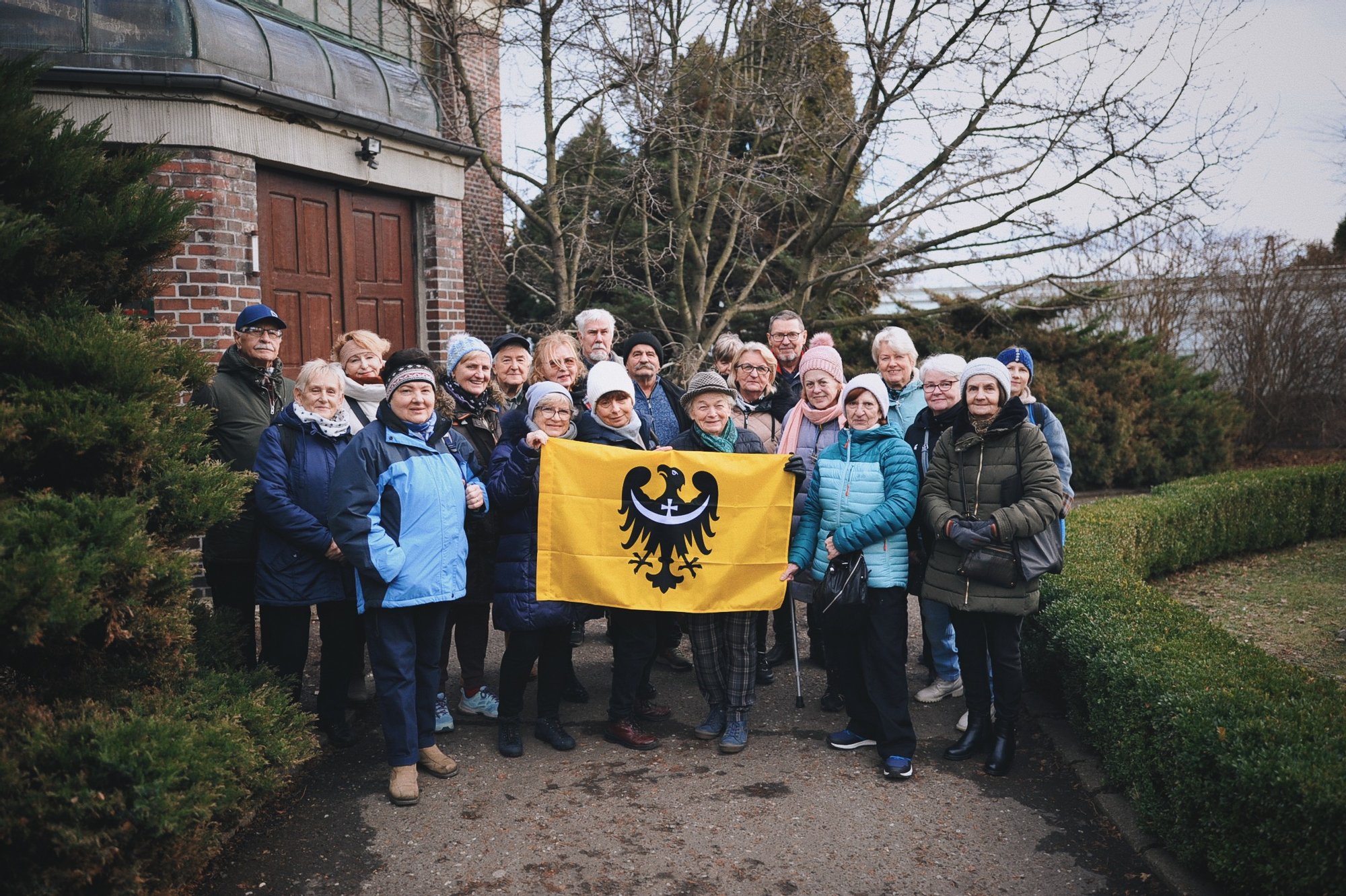 Kolorowa, pozioma fotografia. Grupa seniorów i seniorek pozuje do zdjęcia z flaga Dolnego Śląska przed palmiarnią w Wałbrzychu.