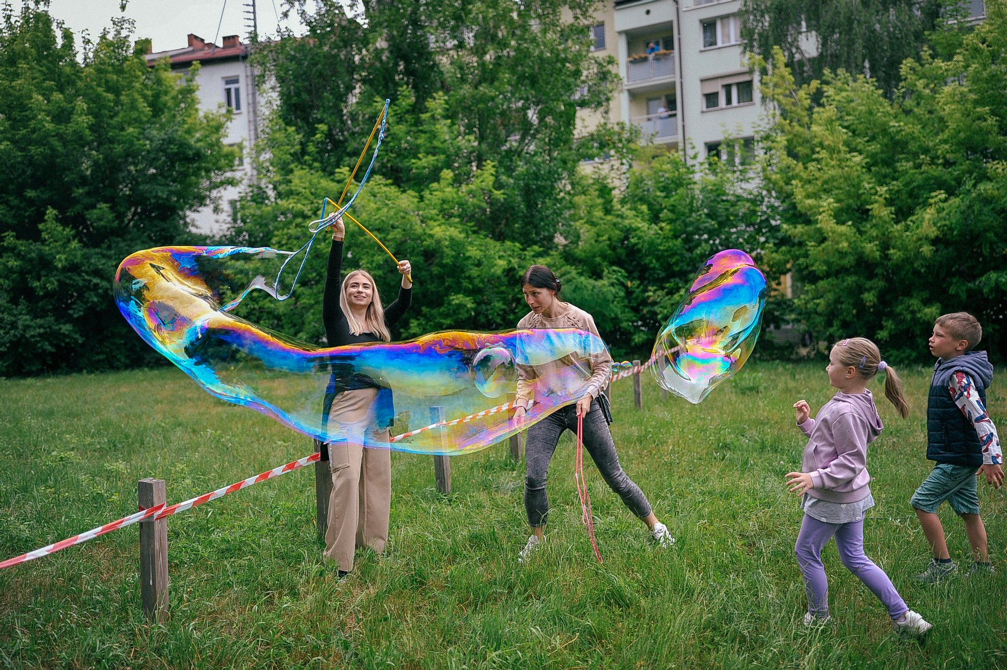 Kolorowa fotografia wykonana podczas święta osiedla. Dzieci i animatorki puszczają na łące ogromne bańki mydlane.