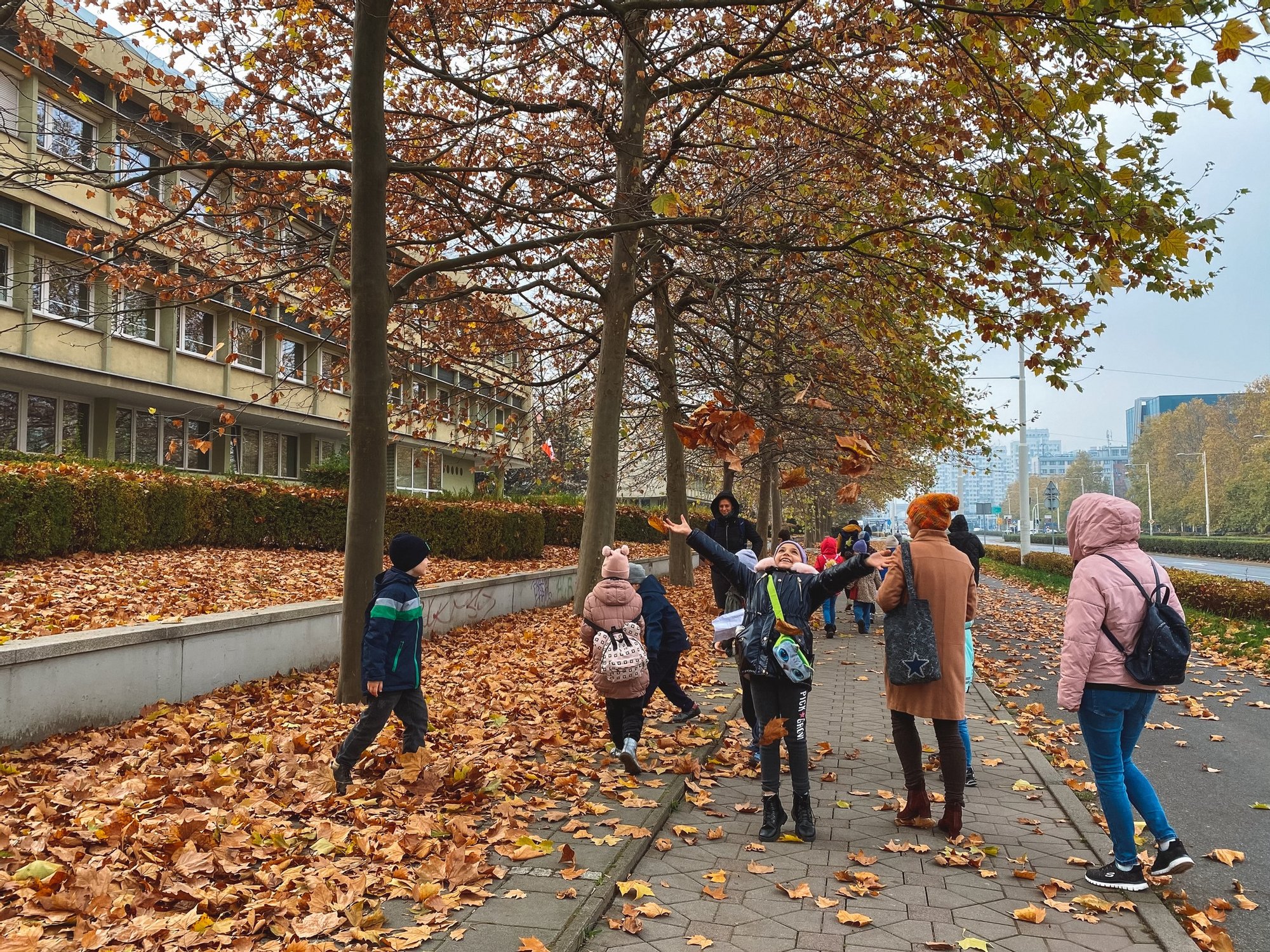 Grupa dzieciaków spaceruje chodnikiem zasypanym jesiennymi liśćmi. Jedna z dziewczynek podrzuca garść liści w powietrze. Trwa spacer po osiedlu.