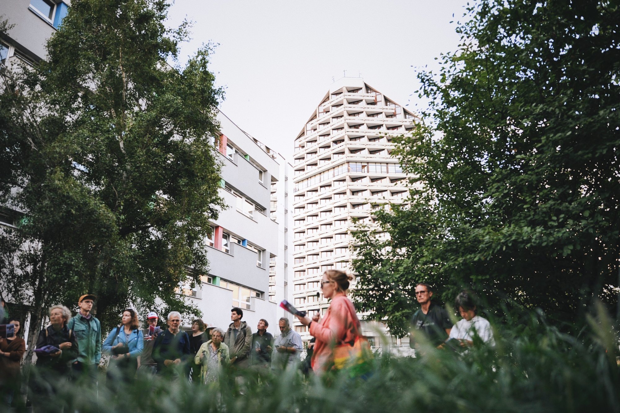 Kolorowa, pozioma fotografia przedstawia grupę osób podczas spaceru z oprowadzaniem po osiedlu. Osoby stoją pod drzewami, w tle za nimi widoczne akademiki Kredka i Ołówek.