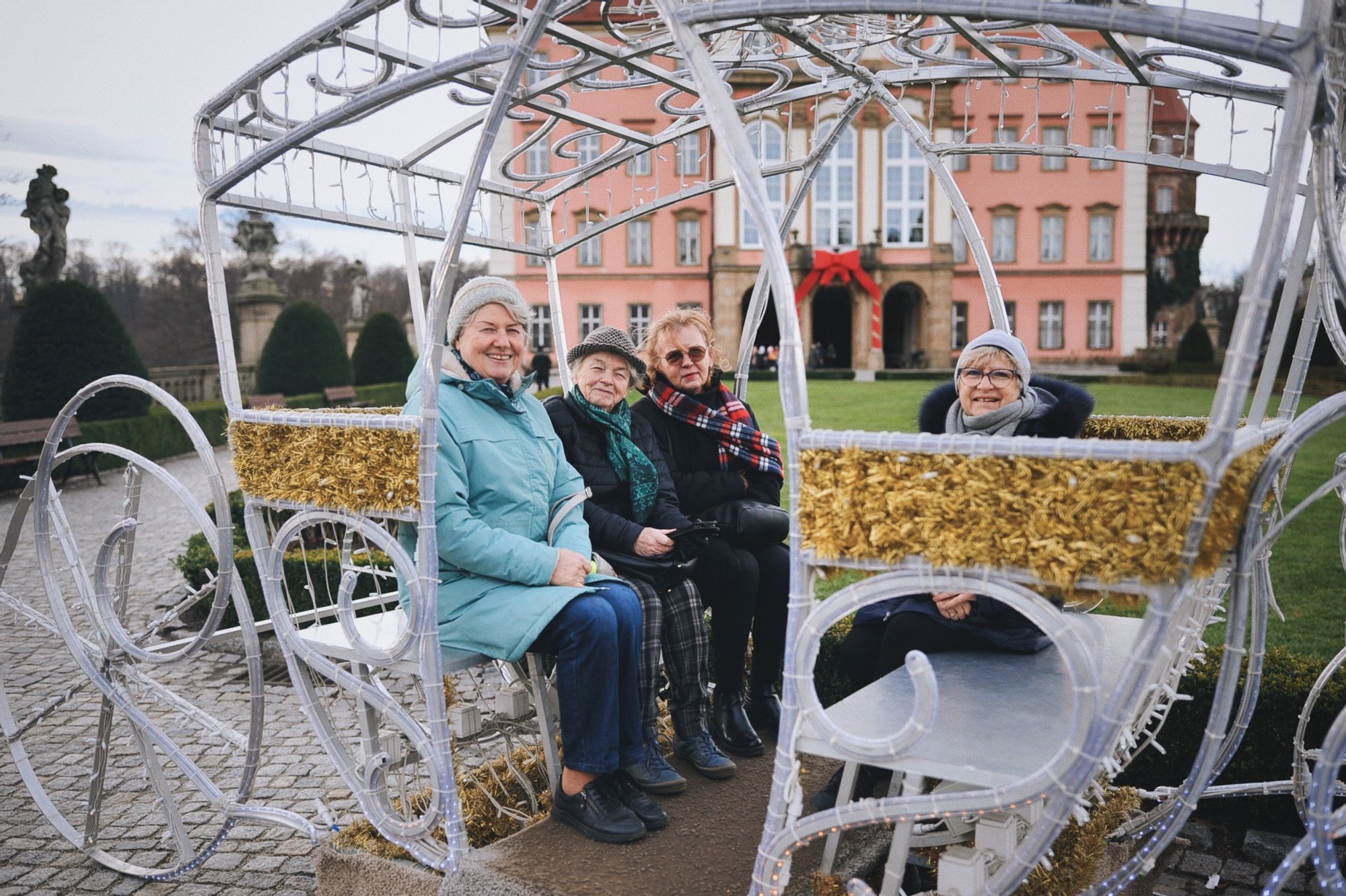 Kolorowa, pozioma fotografia: cztery uśmiechnięte seniorki pozują do zdjęcia w ażurowej karocy przed zamkiem w Wałbrzychu.