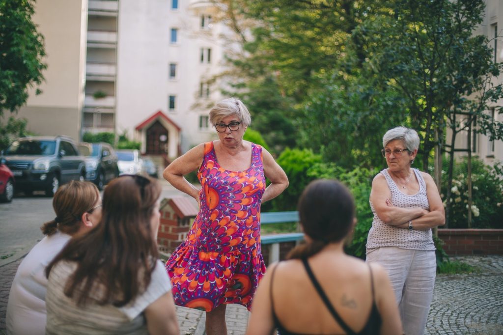 Kolorowa, pozioma fotografia: grupa kobiet zebrała się na podwórku. Kobieta na środku zdjęcia ma na sobie kolorową sukienkę, a kobieta po prawej stronie ma skrzyżowane ręce i jest ubrana na biało. Na pierwszym planie po lewej stronie kadru siedzą trzy inne kobiety, które wydają się być młodsze, są zwrócone w stronę starszych kobiet i rozmawiają z nimi.
