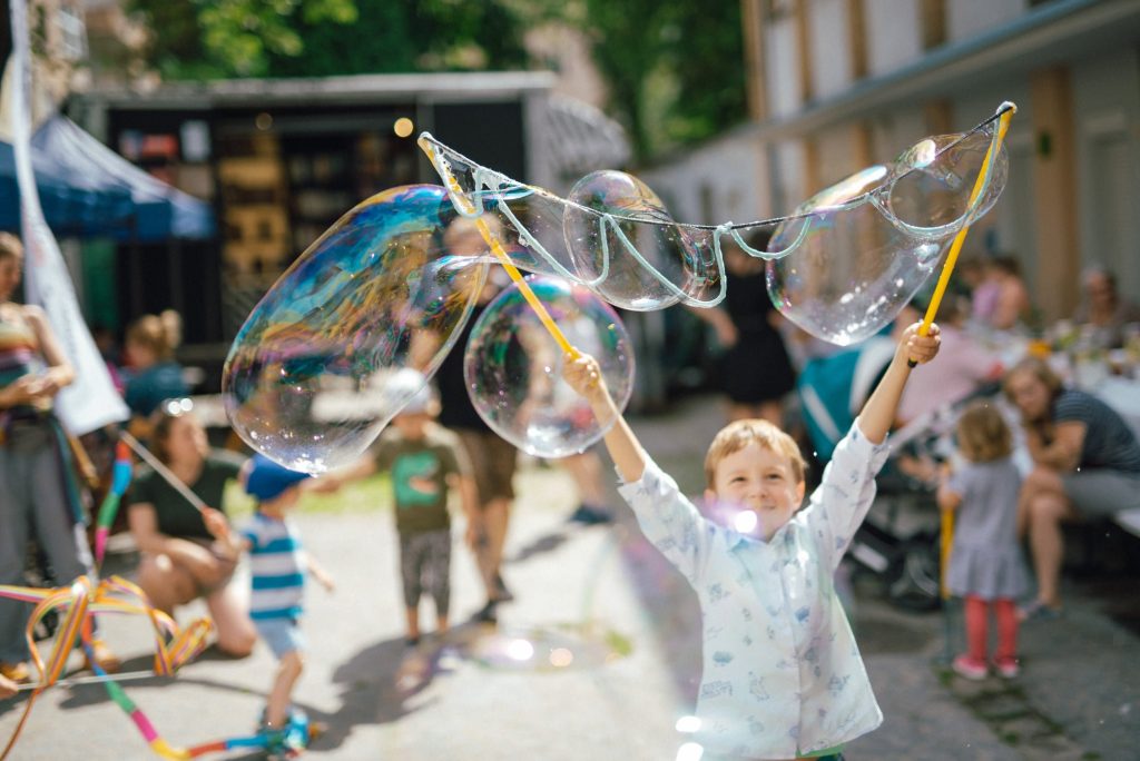 zdjęcie: Marcin Szczygieł Kolorowa, pozioma fotografia przedstawia roześmianego, kilkuletniego chłopca trzymającego w rękach kije do puszczania wielkich baniek mydlanych. Przed nim unosi się kilka takich baniek. W tle za nim widać inne dzieci i osoby dorosłe. Trwa sąsiedzki piknik.