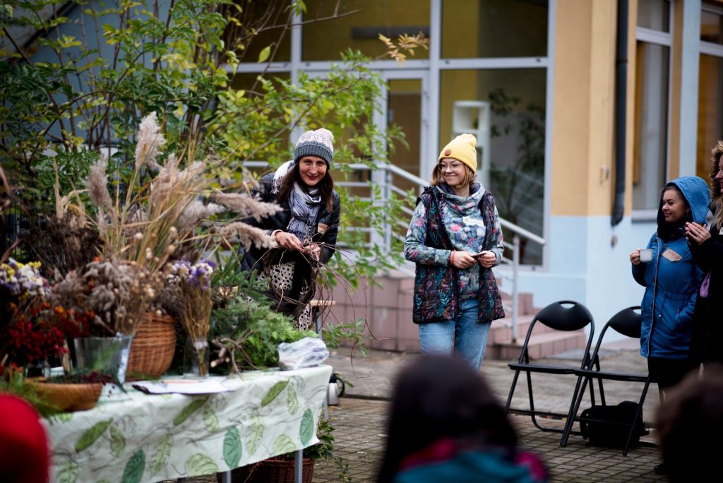 Kolorowa, pozioma fotografia: dwie młode kobiety stoją wokół stołu ustawionego na podwórku. Obok kobiet na stole znajdują się różnego rodzaju rośliny i kwietne bukiety.