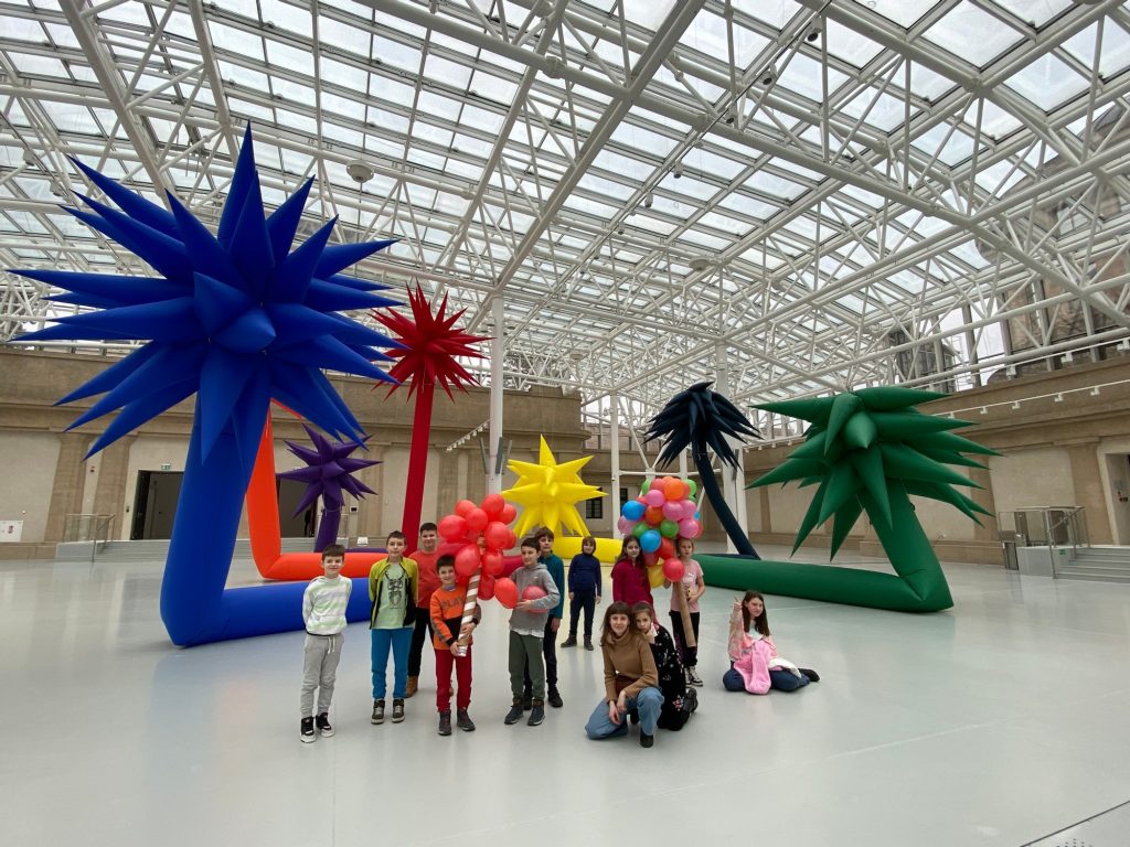 Kolorowa, pozioma fotografia. Grupa dzieci w różnym wieku wraz z opiekunką pozują do zdjęcia w dużej, jasnej muzealnej sali. Za nimi kolorowe dmuchane rzeźby. Kolorowa, pozioma fotografia. Grupa dzieci w różnym wieku wraz z opiekunką pozują do zdjęcia w dużej, jasnej muzealnej sali. Za nimi kolorowe dmuchane rzeźby.