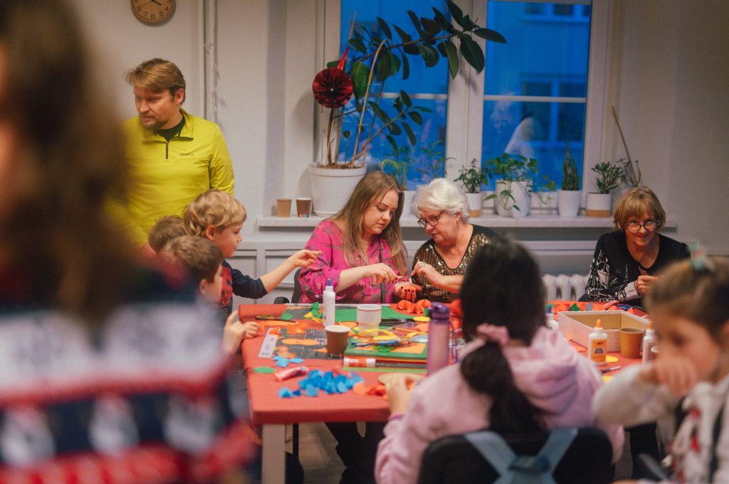 zdjęcie: Marcin Szczygieł Kolorowa, pozioma fotografia: w jasnym pomieszczeniu przy dużym stole siedzi grupa osób w różnym wieku, zarówno dzieci, jak i seniorek. Starsza kobieta razem z młodą dziewczyną kleją razem ozdoby choinkowe. Na stole porozkładanych jest dużo materiałów plastycznych. Dookoła stołu kręcą się inni ludzie. Trwa sąsiedzkie spotkanie świąteczne.