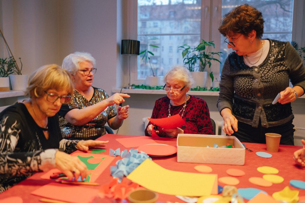 Kolorowa, pozioma fotografia: w jasnym pomieszczeniu przy stole siedzą cztery seniorki, przed nimi rozłożone są materiały plastyczne. Kobiety robią ozdoby świąteczne.