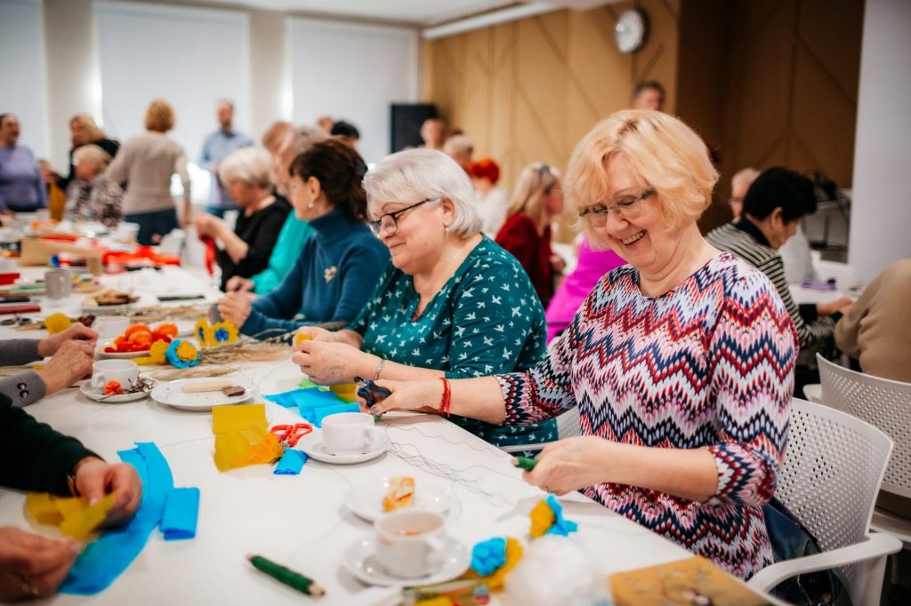 Kolorowa fotografia przedstawia grupę seniorów i seniorek siedzących przy długim stole w jasnym pomieszczeniu. Na stole porozkładane są talerzyki z przekąskami oraz filiżanki z kawą, a także materiały plastyczne. Osoby przy stole wykonują kwiaty z bibuły. Trwa spotkanie międzykulturowej kawiarenki.
