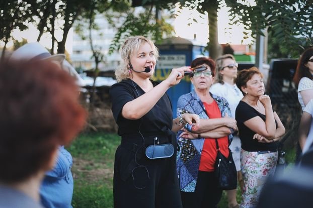 Kolorowa, pozioma fotografia przedstawiająca osoby na spacerze z po osiedlu. Pośrodku zdjęcia prowadząca spacer. Przy ustach ma mikrofon. Wokół niej osoby w różnym wieku.