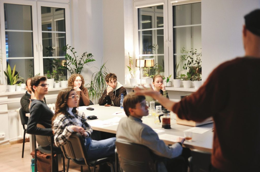 zdjęcie: Anna Pazdej Kolorowa, pozioma fotografia: w jasnym pomieszczeniu przy dużym stole siedzi grupa młodych osób, wszyscy patrzą na ekran znajdujący się poza kadrem. Przy stole stoi mężczyzna i gestykulując opowiada o czymś.